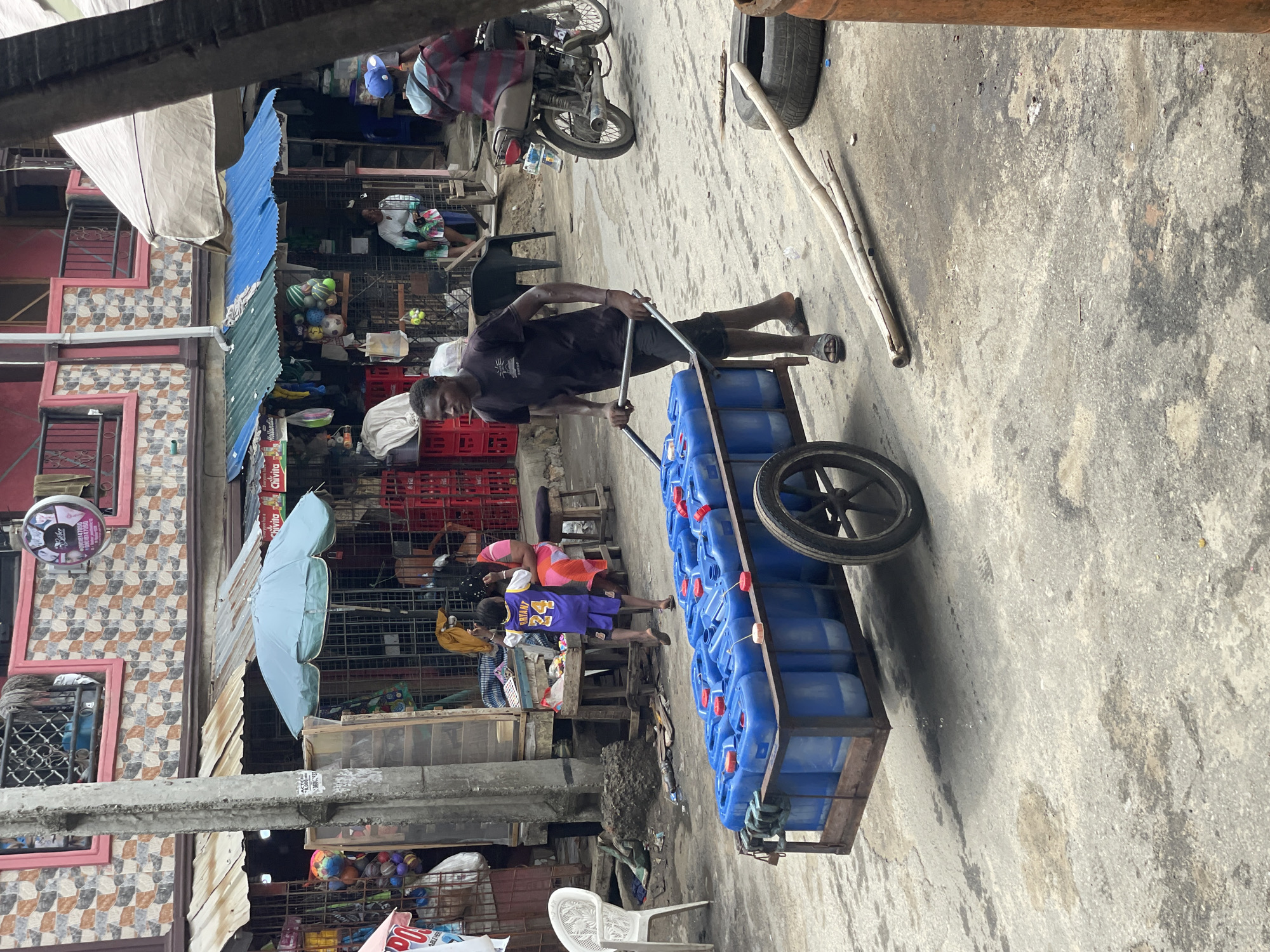 A man is pulling a handcart full of blue jerrycans through a street market in Lagos, Nigeria. The market scene includes shops with corrugated metal roofs, red plastic crates, an umbrella, and people, including a child wearing a Kobe Bryant jersey. A motorcycle is parked in the background, and assorted debris litters the ground. The pavement is cracked and stained.