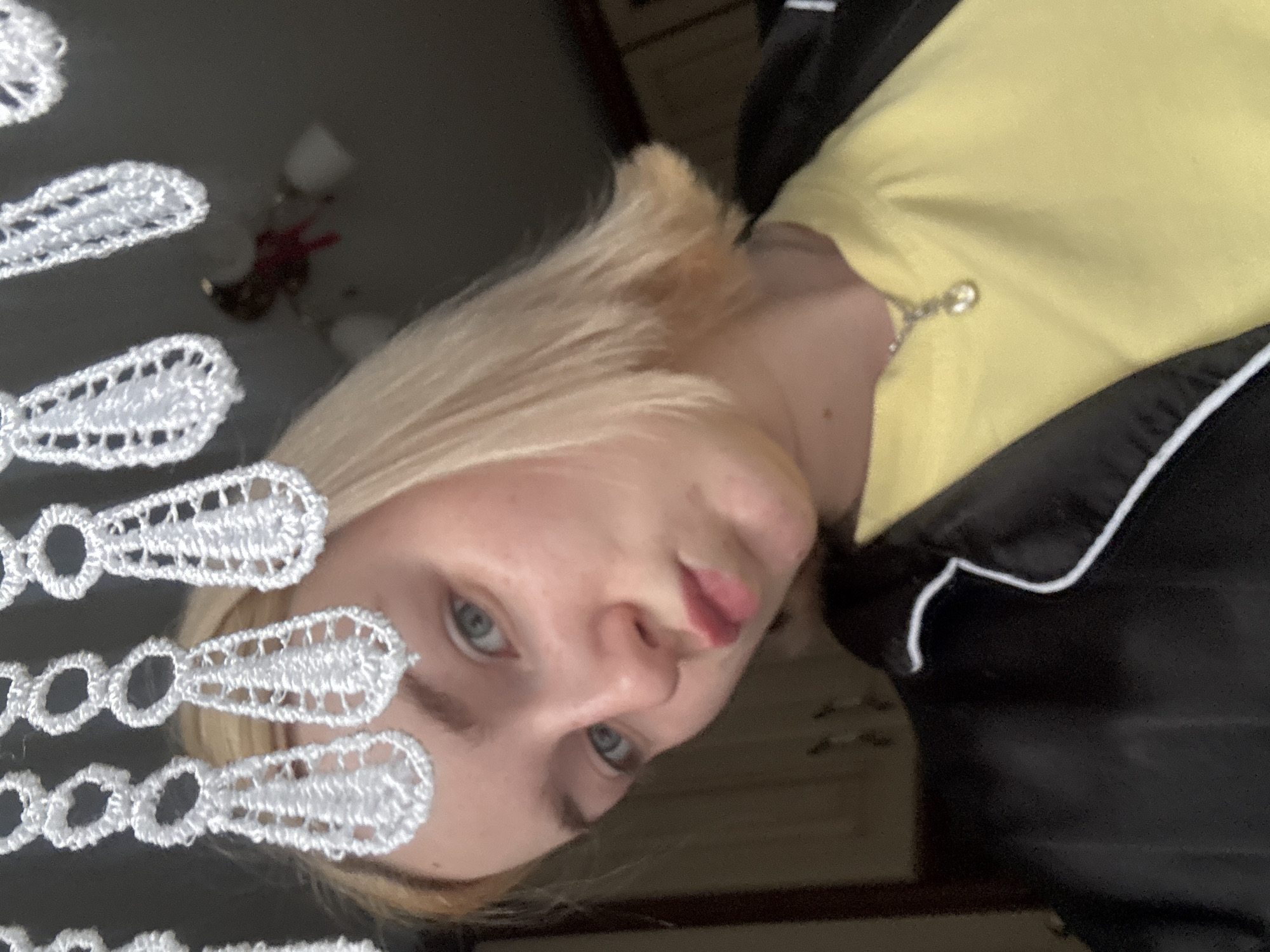 A young blonde woman with blue eyes and fair skin is looking directly at the camera, making a kissy face. She is wearing a yellow t-shirt and a black jacket with white trim. A delicate silver necklace with a small pendant is visible around her neck. Her hair is cut into a bob that reaches her jawline. In the foreground, a white lace curtain with intricate circular and teardrop patterns hangs above her. The background is dark and out of focus, with a hint of what appears to be kitchen cabinets and a ceiling light fixture, suggesting an indoor setting. The image was taken in Sinyavino, Russia.