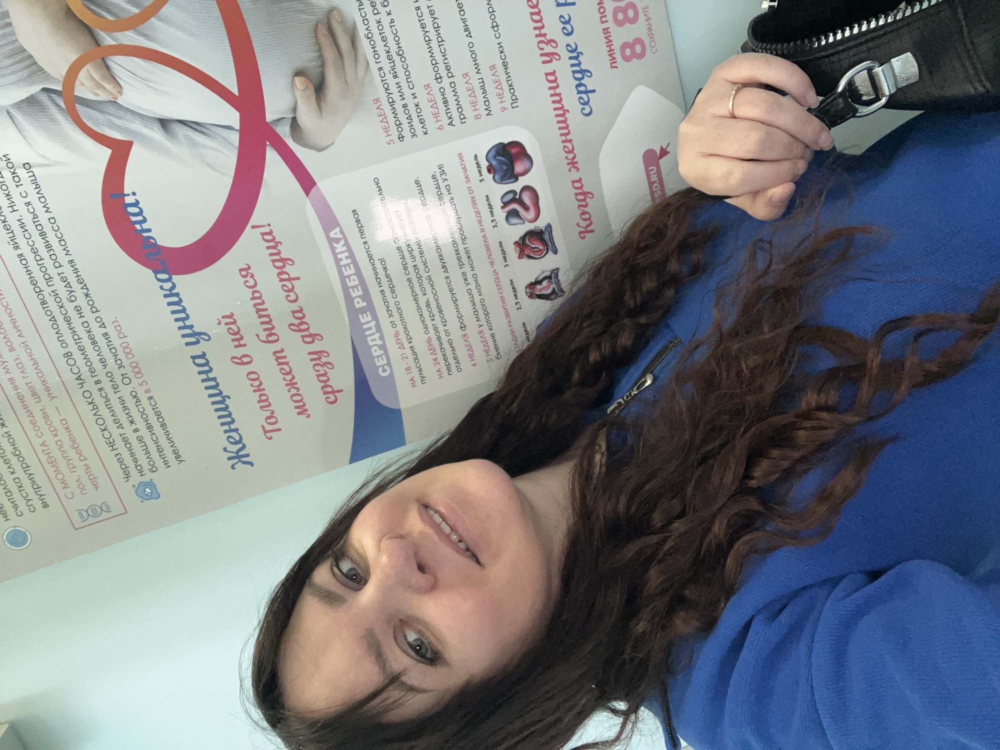 A woman with long, dark, wavy hair is taking a selfie in front of a poster written in Russian about fetal heart development. The poster features a graphic of a pregnant woman and stages of heart development from 2.5 to 5 weeks. Text on the poster includes phrases such as "A woman is unique! Only in her can beat two hearts at once!" and "Heart of the child." The woman is wearing a blue sweatshirt and a gold ring on her finger. She is holding a black purse in her hand. The location is most likely in Noviy, Russia, based on the ".SO.RU" URL shown in the image.