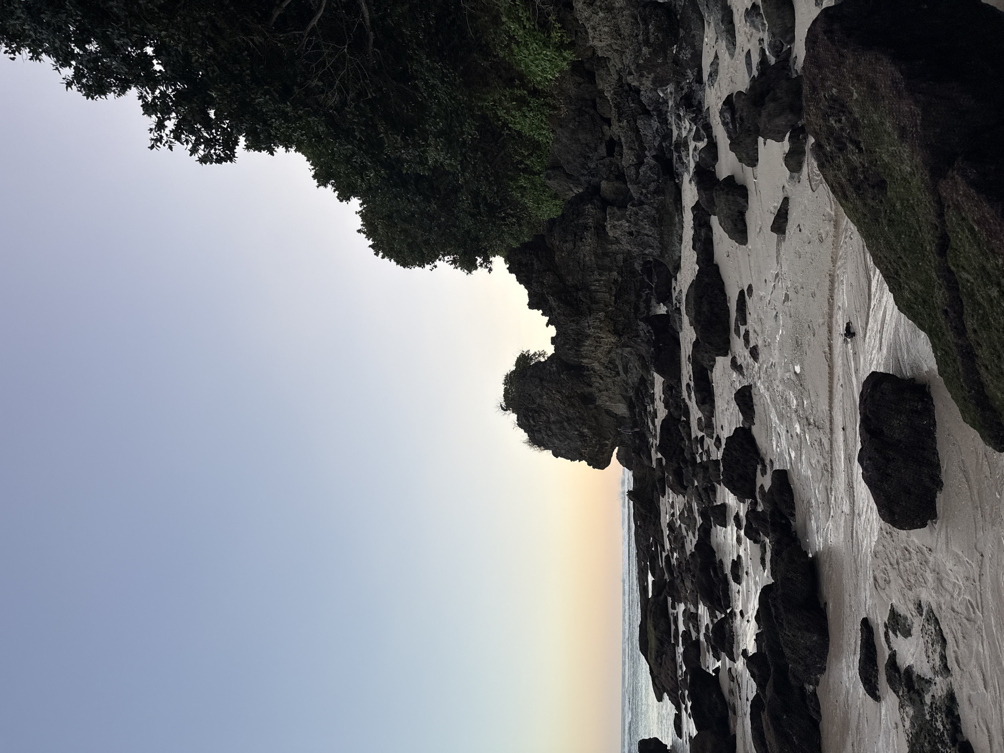 The image depicts a beach scene with dark, wet rocks scattered across a sandy shore. The ocean is visible in the background, with gentle waves breaking. Lush green trees and vegetation cling to a rocky cliff that rises on the right side of the frame. The sky is a gradient of pale blue and soft yellow, suggesting either dawn or dusk. The overall impression is one of a rugged, natural coastline. This scene is located in Kuta Selatan, Indonesia.