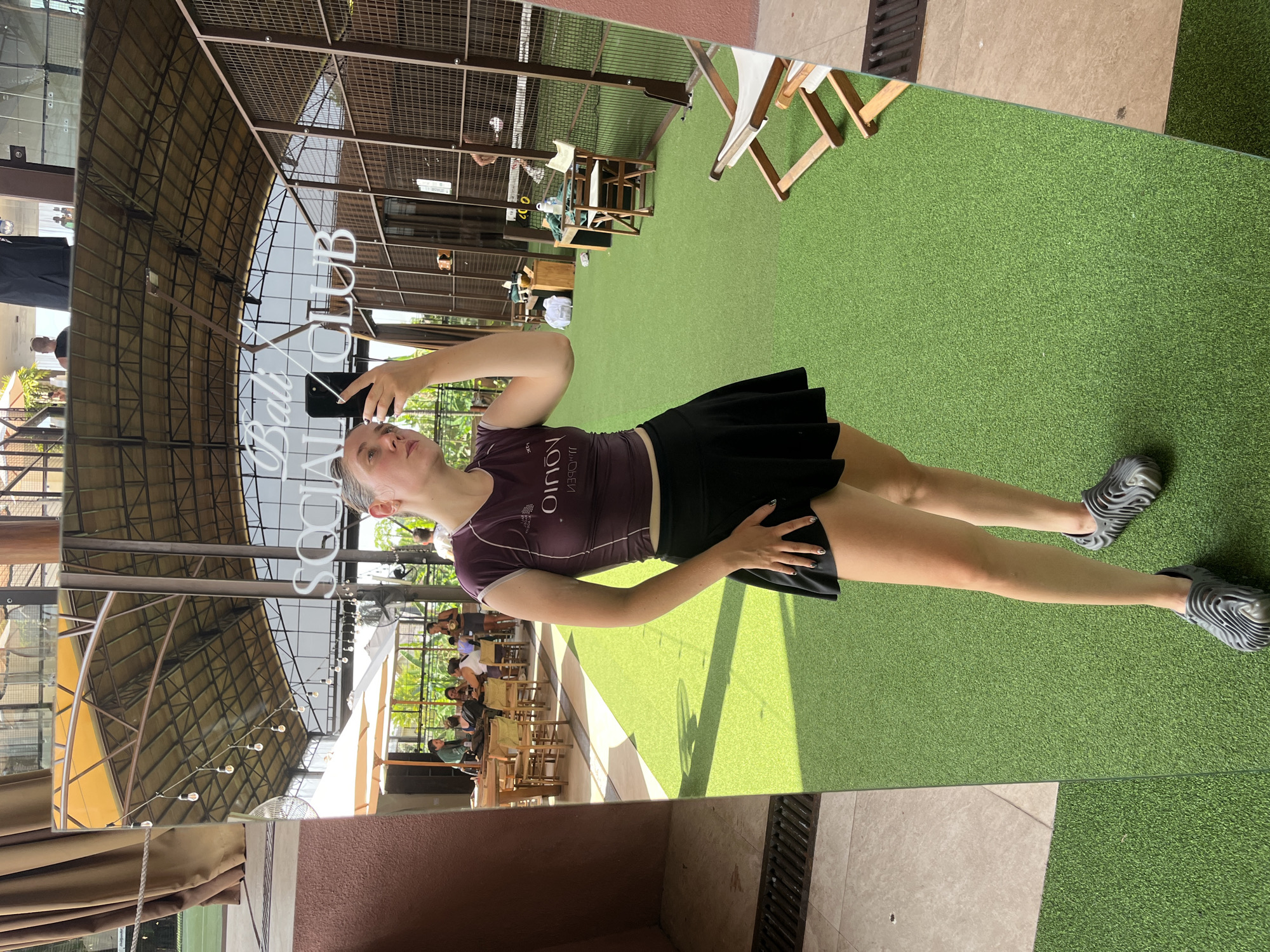 A fair-skinned person is taking a selfie in a large, standing mirror, reflecting an outdoor seating area. The person is wearing a short-sleeved, dark purple top and a black skirt, with gray, patterned shoes. They are holding a cell phone up and looking at the screen. The outdoor seating area features wooden tables and chairs under a large, open-air structure. The ground is covered in artificial green turf. The mirror also reflects the words "Social Bali Club". The location appears to be in North Kuta, Indonesia.