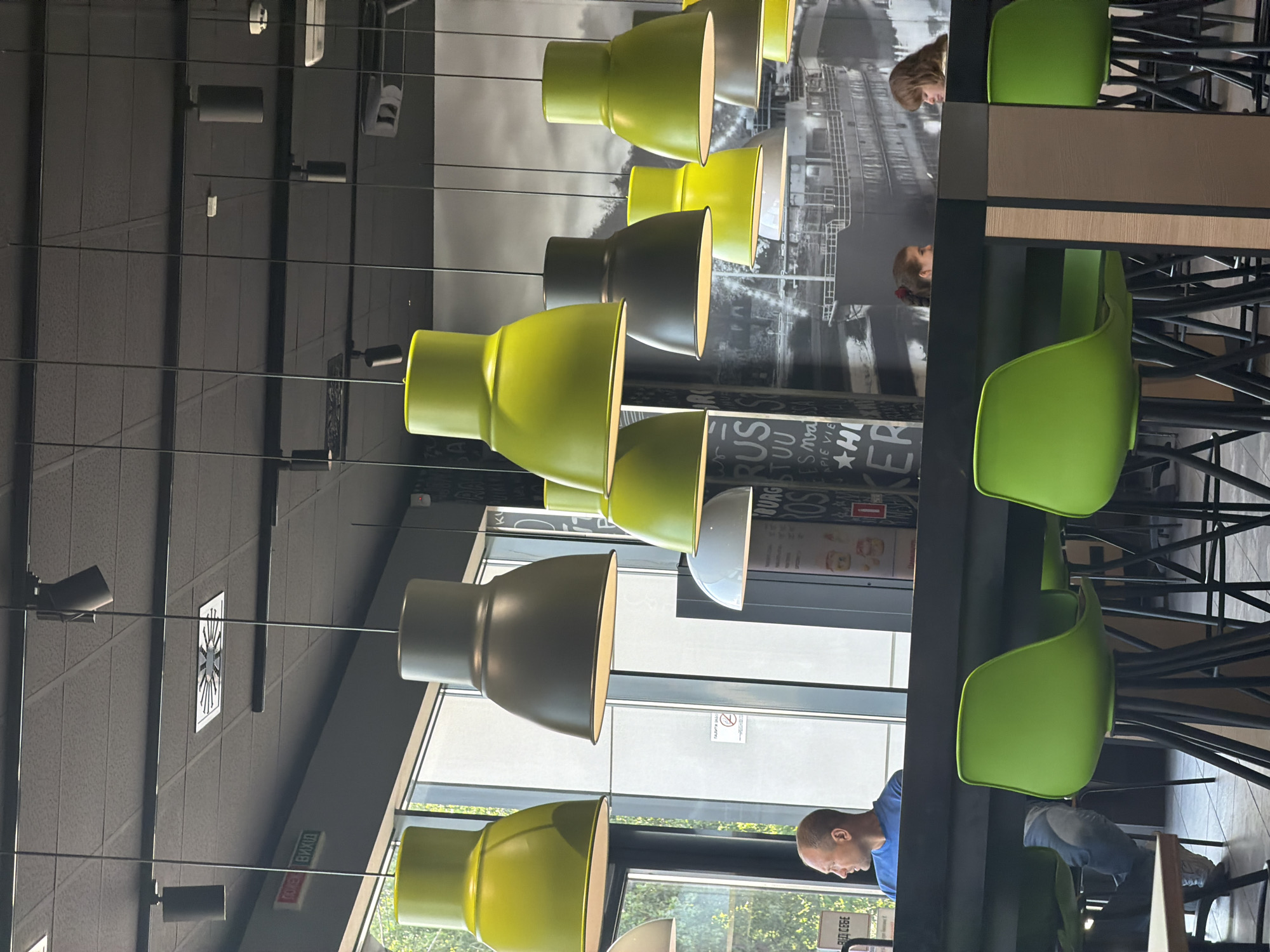 The image depicts the interior of a McDonald's restaurant in Kyiv, Ukraine. The scene is a bright and modern dining area, characterized by its high ceiling with black acoustic tiles and an array of pendant lights. Several of these lights have lime green shades, while others are a dark gray or white.
In the foreground, a long, dark counter runs horizontally across the image, separating the seating area. To the left of the counter, a man wearing a blue t-shirt and jeans is seated and looking to his left. In the background, several lime green bar stools with black legs are visible.
Further back, a large black-and-white mural adorns the wall, featuring images of a ship and text. Two young women are seated at the counter in the background, facing each other and seemingly engaged in conversation. To the left of the man in the blue t-shirt, a window with green foliage visible outside suggests the restaurant is located in an area with trees.
On the wall above the counter, a red and green sign with white Cyrillic lettering is visible, reading "ПОЖЕЖНА ВИХІД" which translates to "FIRE EXIT." The overall ambiance suggests a casual dining setting within a well-lit establishment.