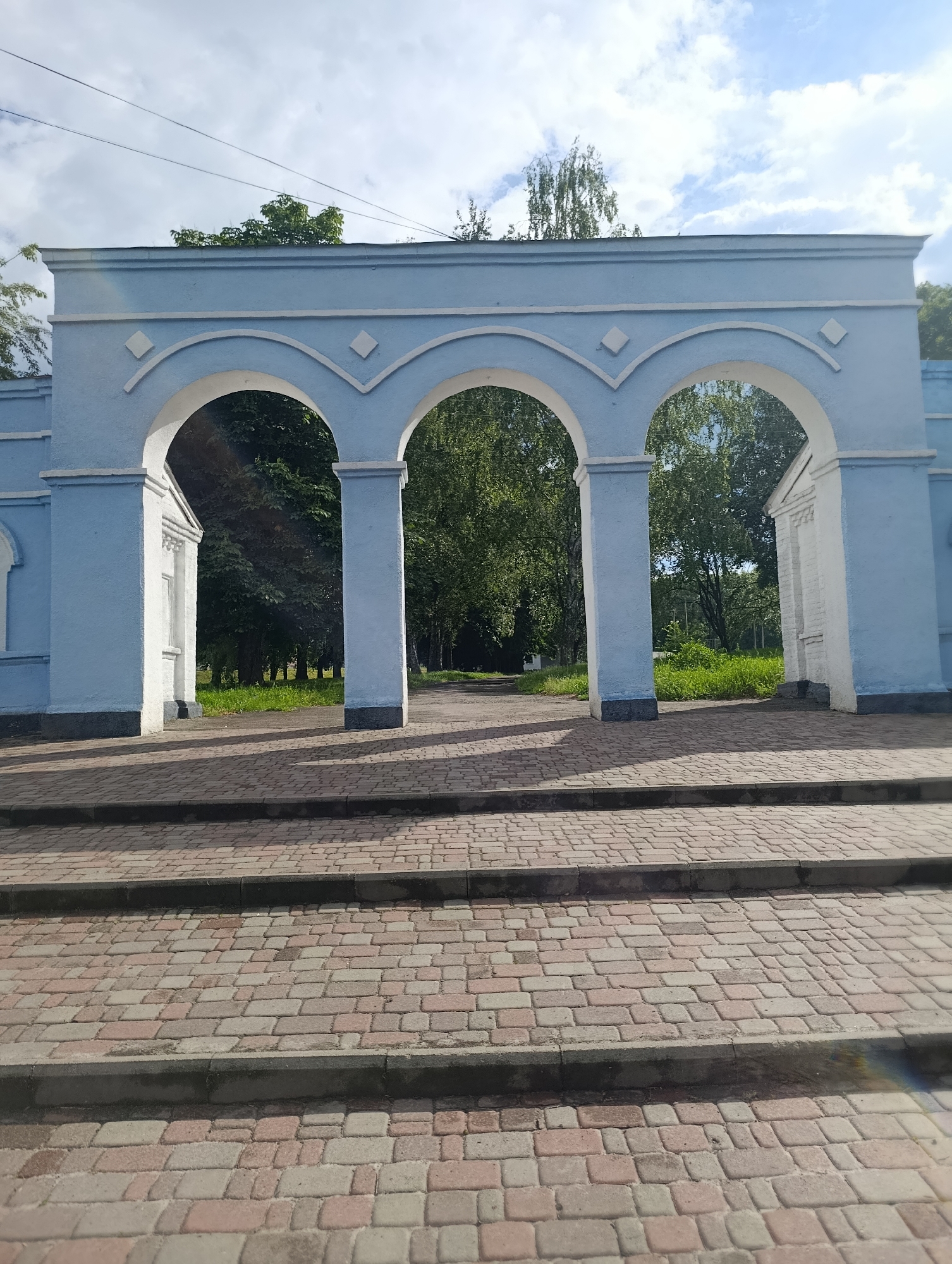 The image shows a light blue archway with three openings, set against a background of trees and a cloudy sky. The archway is constructed of concrete and painted white and blue, with decorative diamond shapes above each arch. In front of the archway is a paved walkway made of interlocking bricks, with three steps leading up to the structure. Beyond the archway, a path leads into a park-like area with lush green grass and trees. The overall impression is one of an entrance to a park or public space. This archway is located in the city of Khorol, Ukraine.