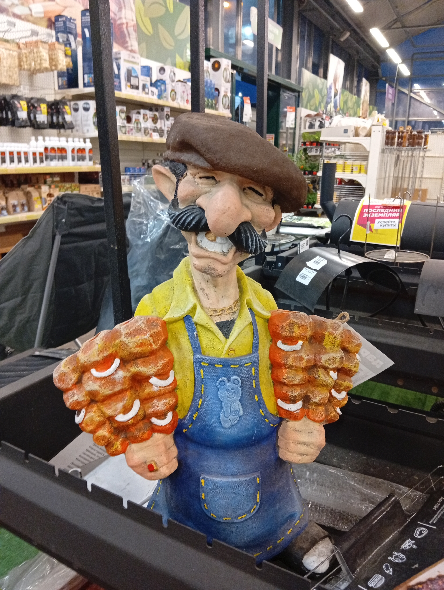 A static, eye-level medium shot captures a decorative garden statue of a caricature of a man wearing a brown beret, a yellow shirt, and blue overalls, holding what appears to be cooked ribs in both hands. The statue is positioned inside a black metal barbecue grill, which is likely part of a retail display. The background suggests an indoor retail environment, possibly a home improvement or garden store, with shelves stocked with various goods visible. A yellow sign with Russian text, partially obscured by the statue, is attached to the grill. The overall lighting is artificial, indicative of an indoor setting. The image does not depict any discernible location in the city of Pushkino, Russia, nor does it show any people or actions.