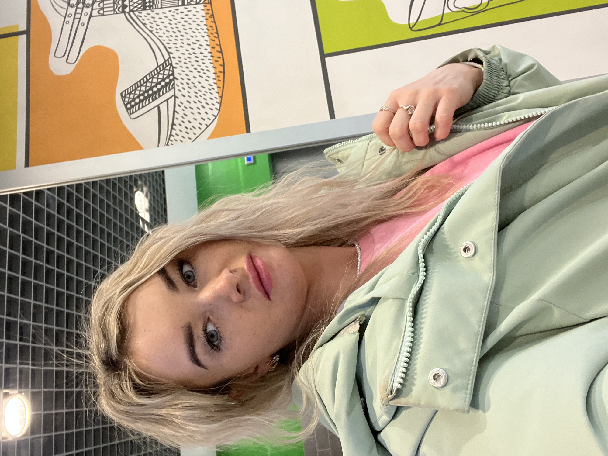 A blonde woman with fair skin is facing the camera, wearing a light green jacket over a pink shirt, with several rings on her fingers. She is in front of a wall with patterned panels and a tiled wall, suggesting an indoor location. A light source is visible to the left. The location is Izhevsk, Russia.