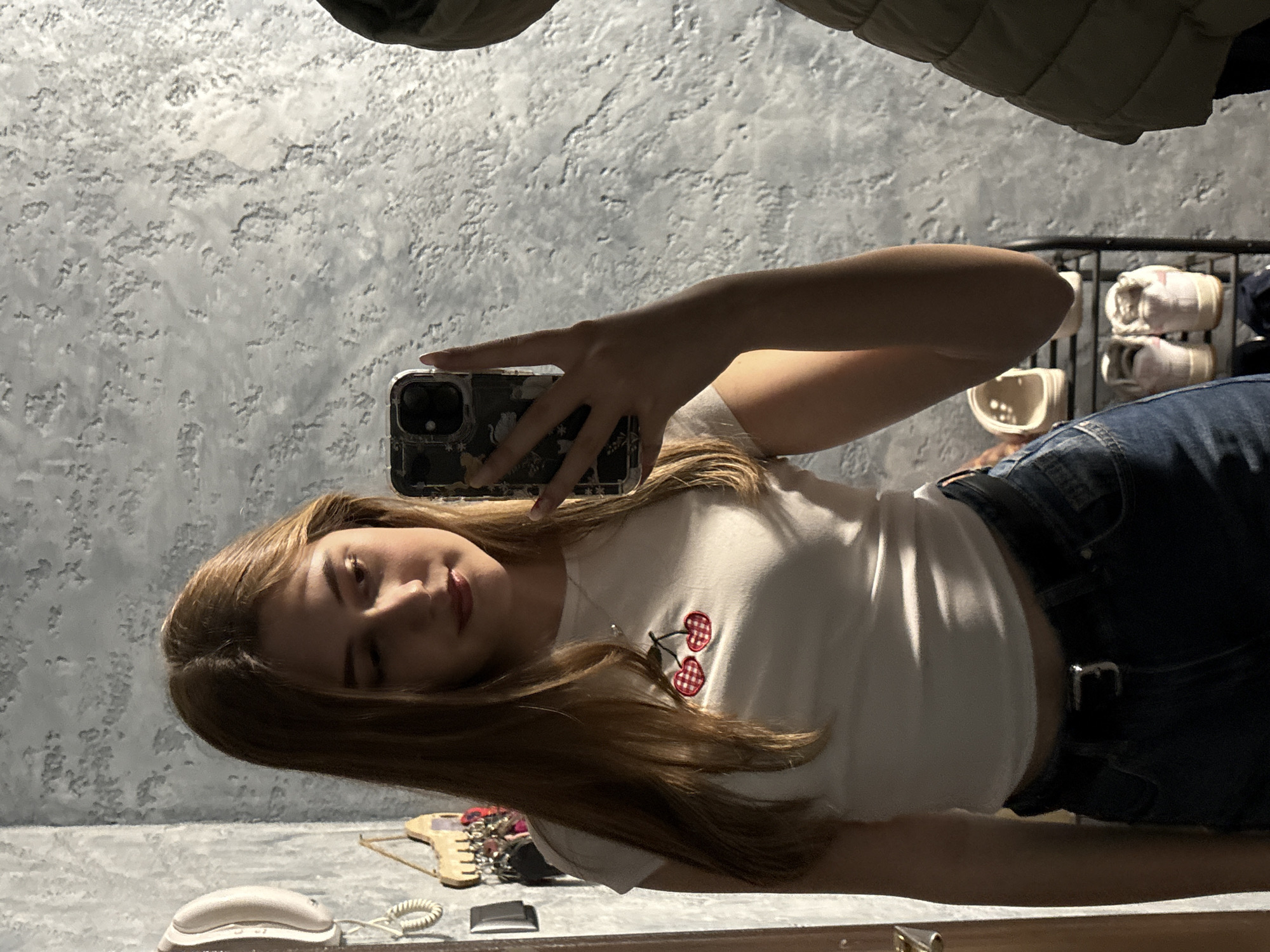 A young woman is taking a selfie in a mirror. She is wearing a white t-shirt with a cherry design, blue jeans, and a black belt. Her hair is long and brown. In the background, there is a textured gray wall and a shelf with shoes. On the surface in front of the mirror, there is a white landline telephone, a small grey object, some keys, and other personal items. There is no information in the image that indicates the location is in the city of Panagyurishte, Bulgaria.
