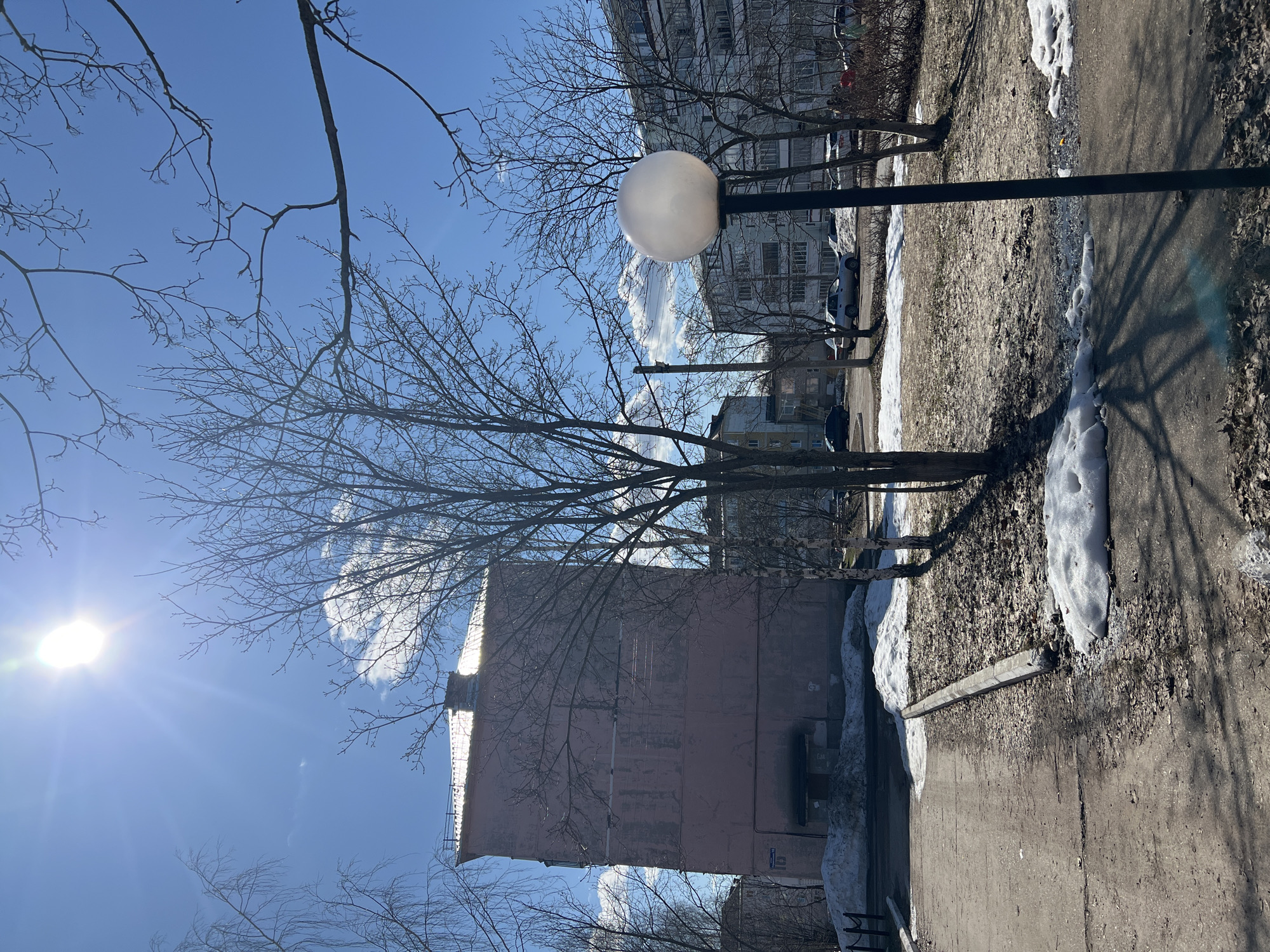The image depicts a street scene in Noviy, Russia. A tall, weathered pink apartment building rises against a bright blue sky with scattered clouds. Bare trees frame the building and sky. A street lamp with a spherical white shade stands near the building. Patches of snow remain along the edge of a sidewalk. A street runs alongside the sidewalk with parked cars visible in the distance. The sun is bright, casting long shadows. No people are visible in the image.