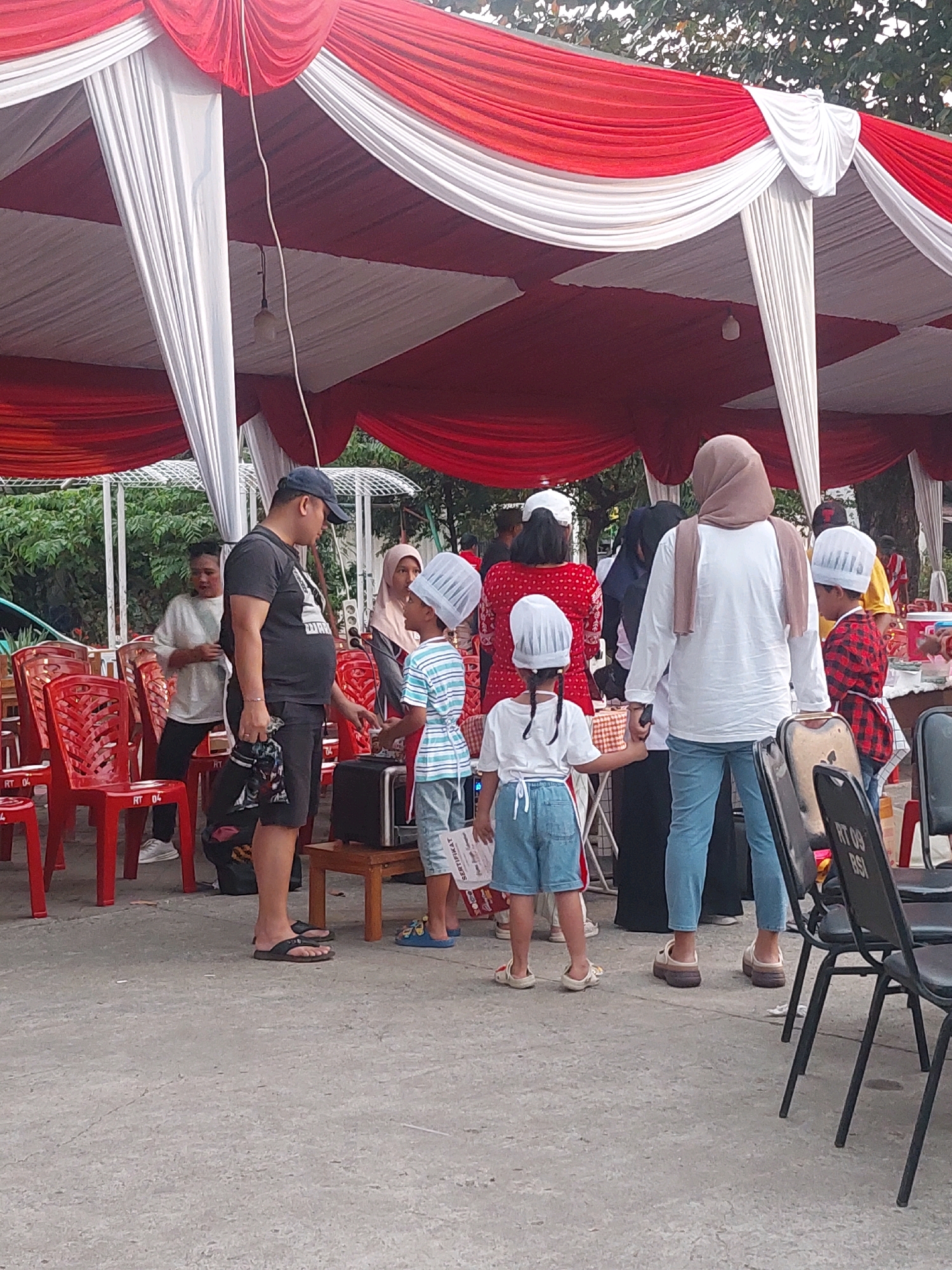 The image depicts an outdoor gathering under a red and white tent, likely in Depok, Indonesia, given the context. A group of people are interacting, with several children wearing chef hats and aprons suggesting a culinary-themed event or activity. In the foreground, a man in a dark t-shirt and shorts is talking to a child in a striped shirt and chef hat. To the right, a woman in a white shirt and headscarf stands near a child in a red checkered shirt and chef hat, who is facing away from the camera. Red plastic chairs are visible throughout the scene, and trees and foliage suggest a park or open outdoor space. The ground is paved with concrete.