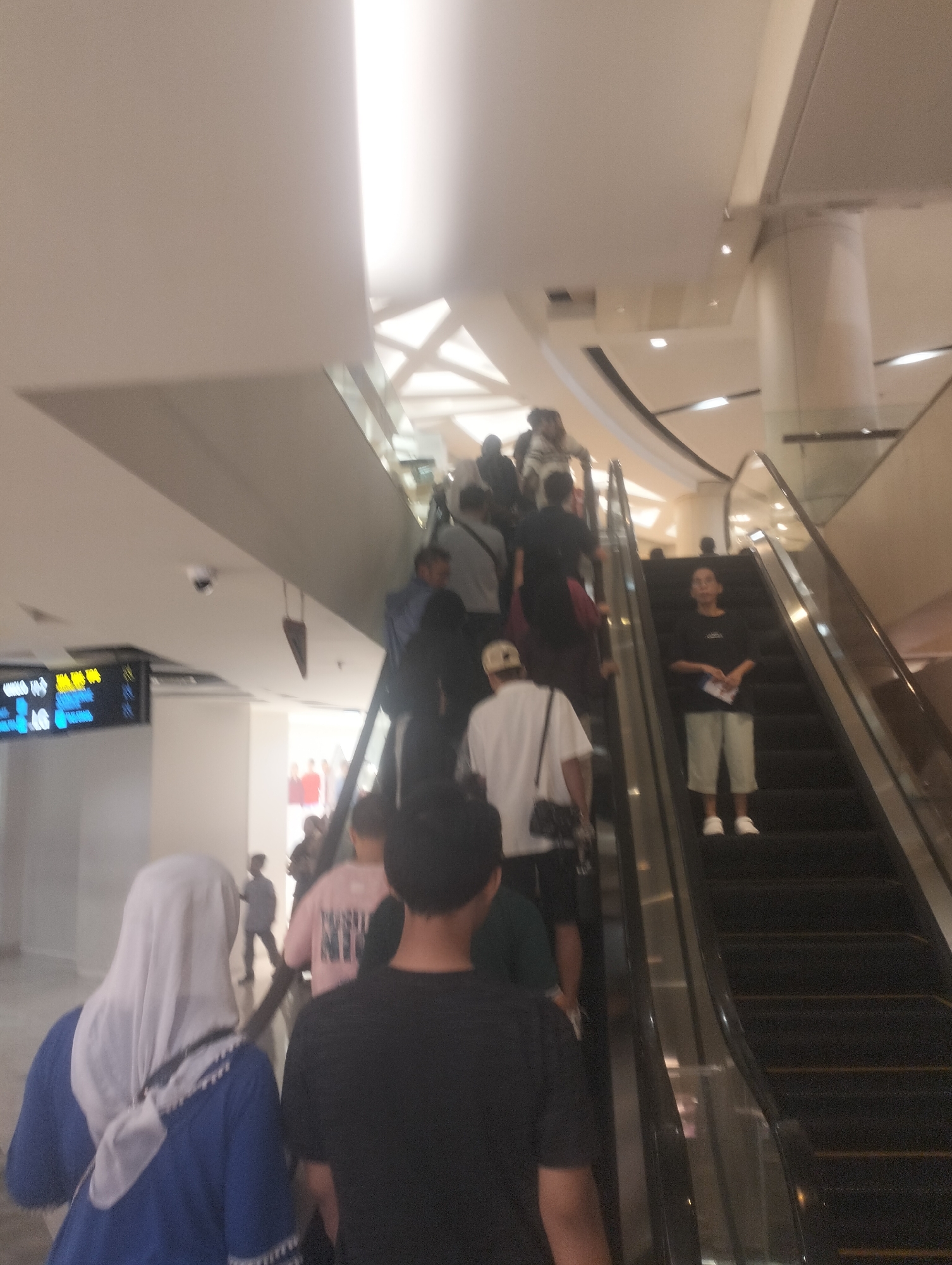 The image depicts an indoor scene, likely a shopping mall or an airport terminal, with a prominent escalator. Several individuals are seen on and around the escalator, moving in both directions. The lighting suggests an indoor environment, with artificial light sources visible in the ceiling. On the left side of the frame, a digital display board shows text and graphics, potentially indicating flight information or store directories, common in locations like the Juanda International Airport in Surabaya, Indonesia. The people are dressed in casual attire, and their faces are largely obscured due to the low resolution and distance. The overall impression is a busy public space with people engaged in transit.