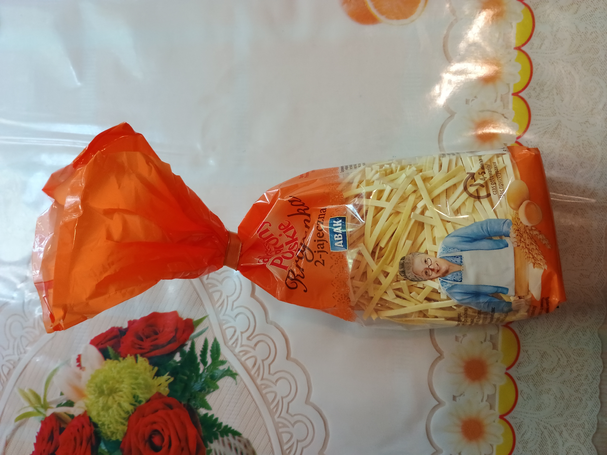 This image shows a bag of Polish egg noodles, possibly "Frytka" brand, standing upright on a white tablecloth. The tablecloth has a decorative lace edge with floral patterns. To the left of the noodle bag, a bouquet of red roses and other flowers is visible. The noodle bag is partially transparent, revealing the yellow noodles inside. The packaging features an illustration of an elderly woman wearing glasses and an apron, along with graphics indicating cooking time and ingredients. The overall setting appears to be a domestic indoor environment. There is no information in the image to indicate its location in the city of Zaporizhzhia, Ukraine.