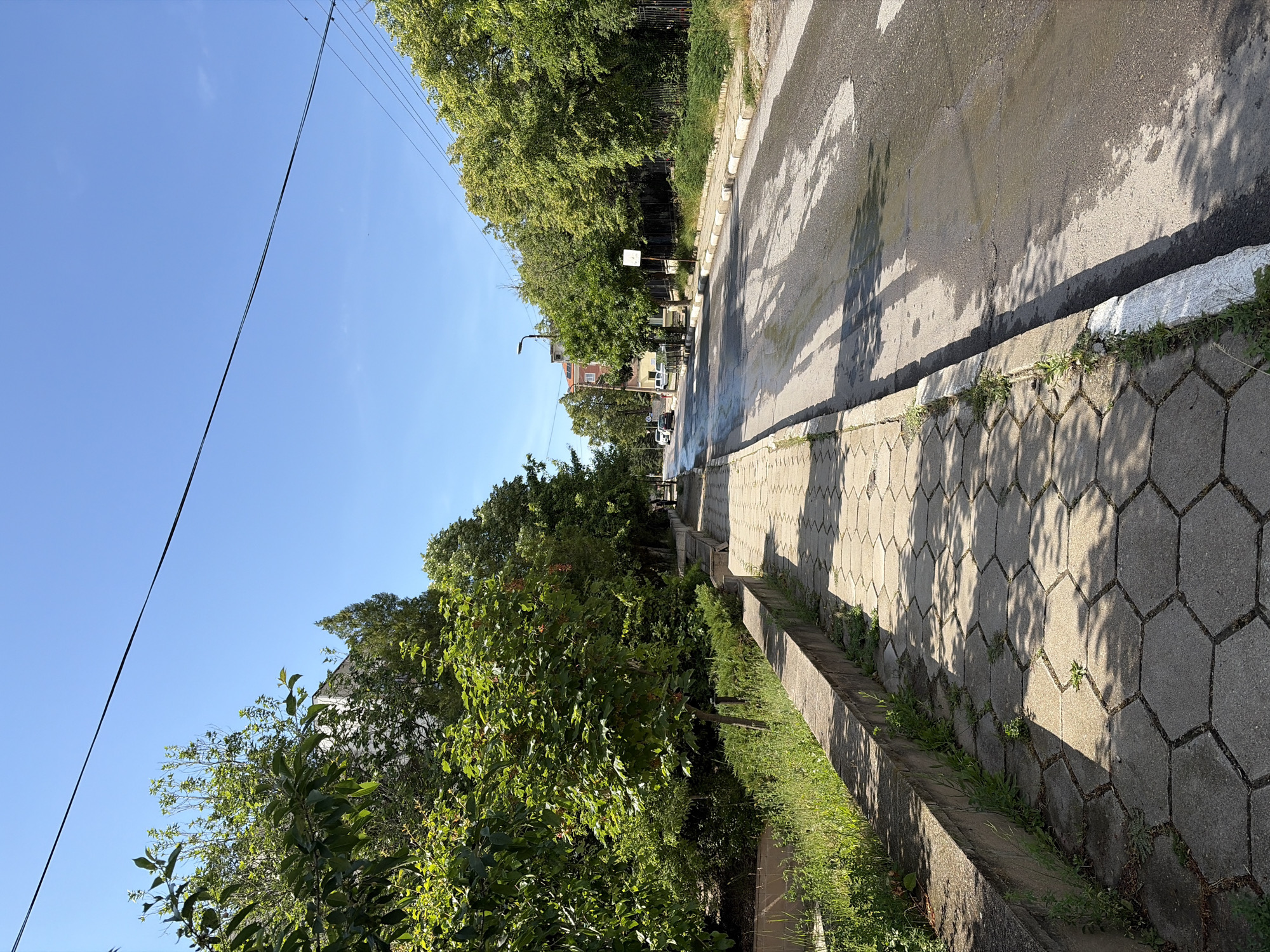 The image shows a street scene in Bobov Dol, Bulgaria. On the right is a patterned concrete sidewalk, bordered by a concrete drainage ditch with grass growing inside. Next to the sidewalk is a wet asphalt road. On the left are trees and foliage, with a white building partially visible through the leaves. In the distance, there are a few cars and buildings. Overhead are power lines against a clear blue sky.
