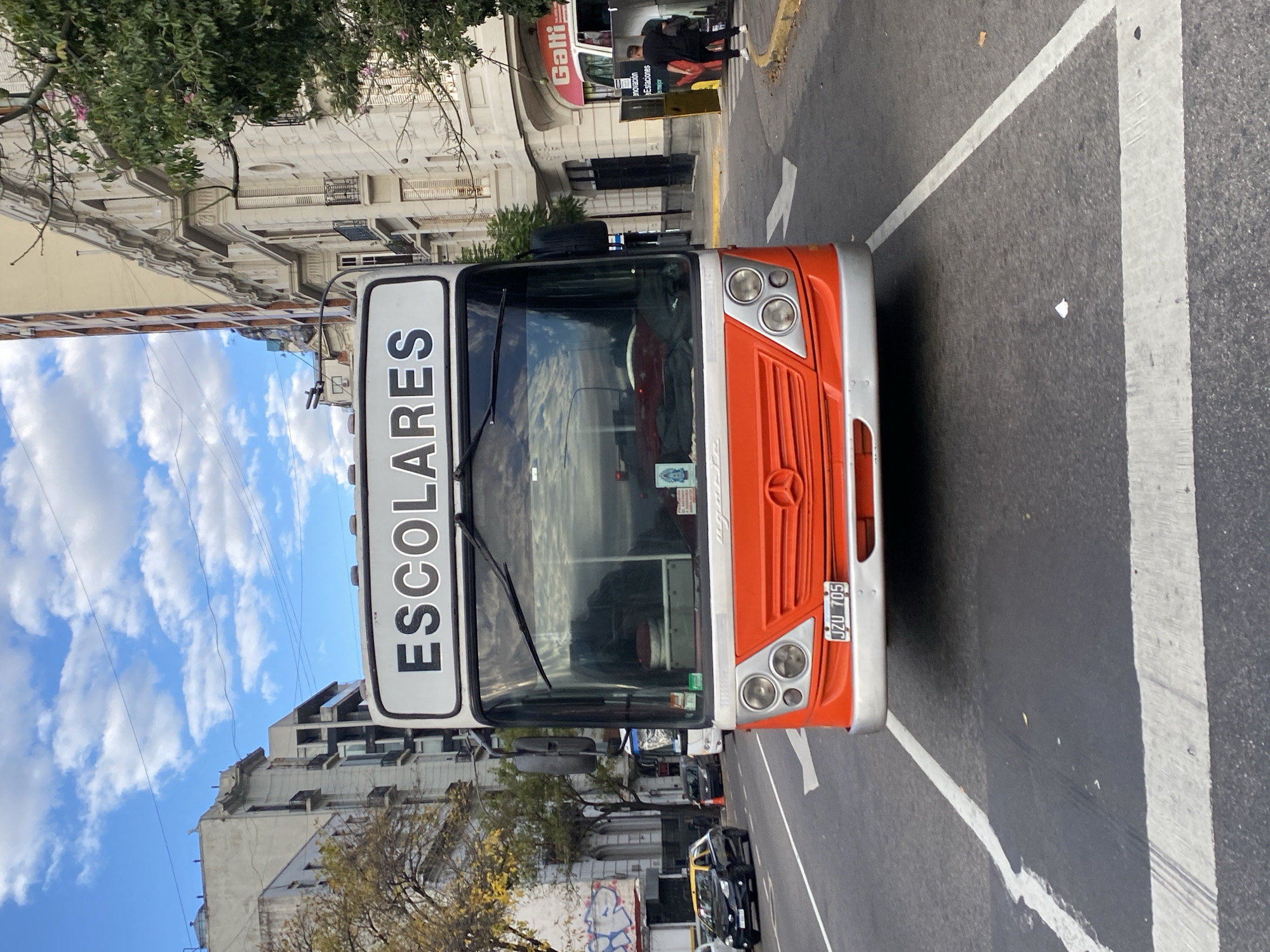 The image shows an orange and silver bus with "ESCOLARES" (scholars) written in bold letters on top of the vehicle. The bus is likely in Buenos Aires, Argentina, based on the vehicle license plate "JZU 705." The street is marked with white crosswalk lines and arrows indicating lane directions. A pedestrian is visible on the sidewalk near a store called "Gatti." Graffiti is visible on a wall. Buildings are of classic design, consistent with the architecture found in Buenos Aires. The sky is blue with fluffy white clouds.