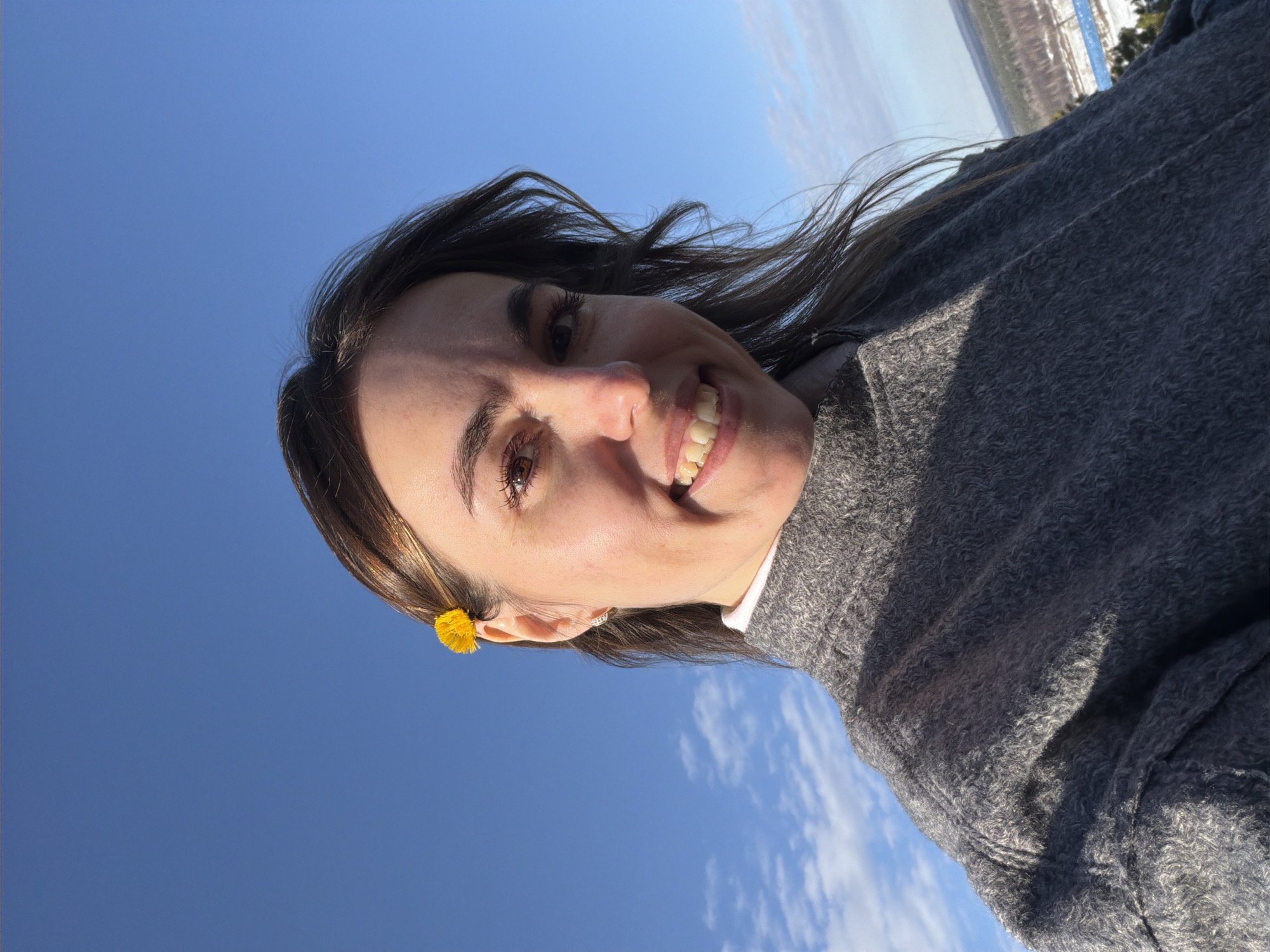 A woman smiles at the camera, angled from below against a clear blue sky. She is wearing a gray sweater and has a yellow flower tucked behind her ear. In the distant background, there is a cityscape. Given the request to include the city of Sarapul, Russia, as the location, the cityscape is likely Sarapul.