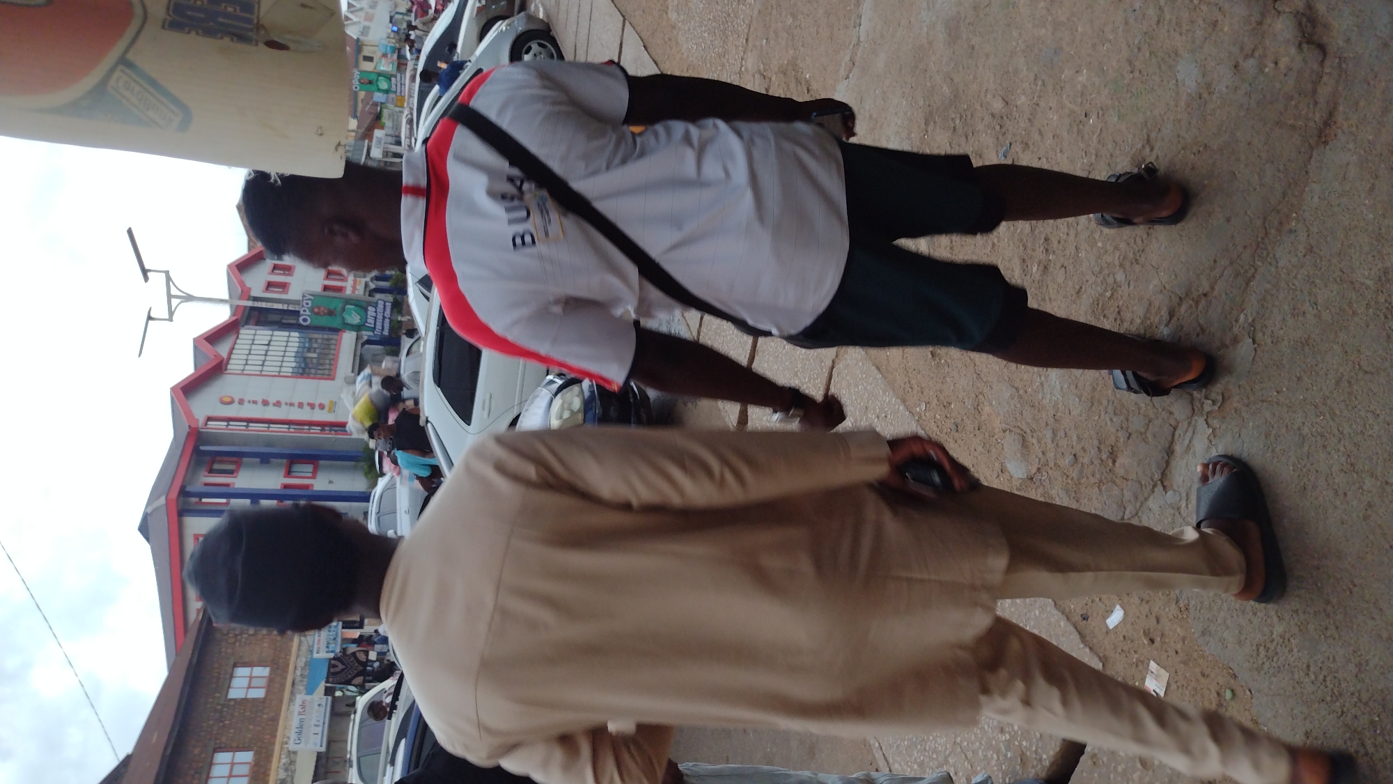 The image captures a street scene in Jos, Nigeria, as evidenced by the visible "Opay" and "Golden Kaily" signage. Two men are in the foreground; one is wearing a white shirt with red accents and dark shorts, along with a black shoulder strap, while the other is wearing a light brown suit and a black cap. Both are wearing sandals. Parked white cars line the street, and a multi-story building with red and white architectural elements is visible in the background. Other pedestrians are present but not clearly defined. The ground appears to be a concrete or paved surface.