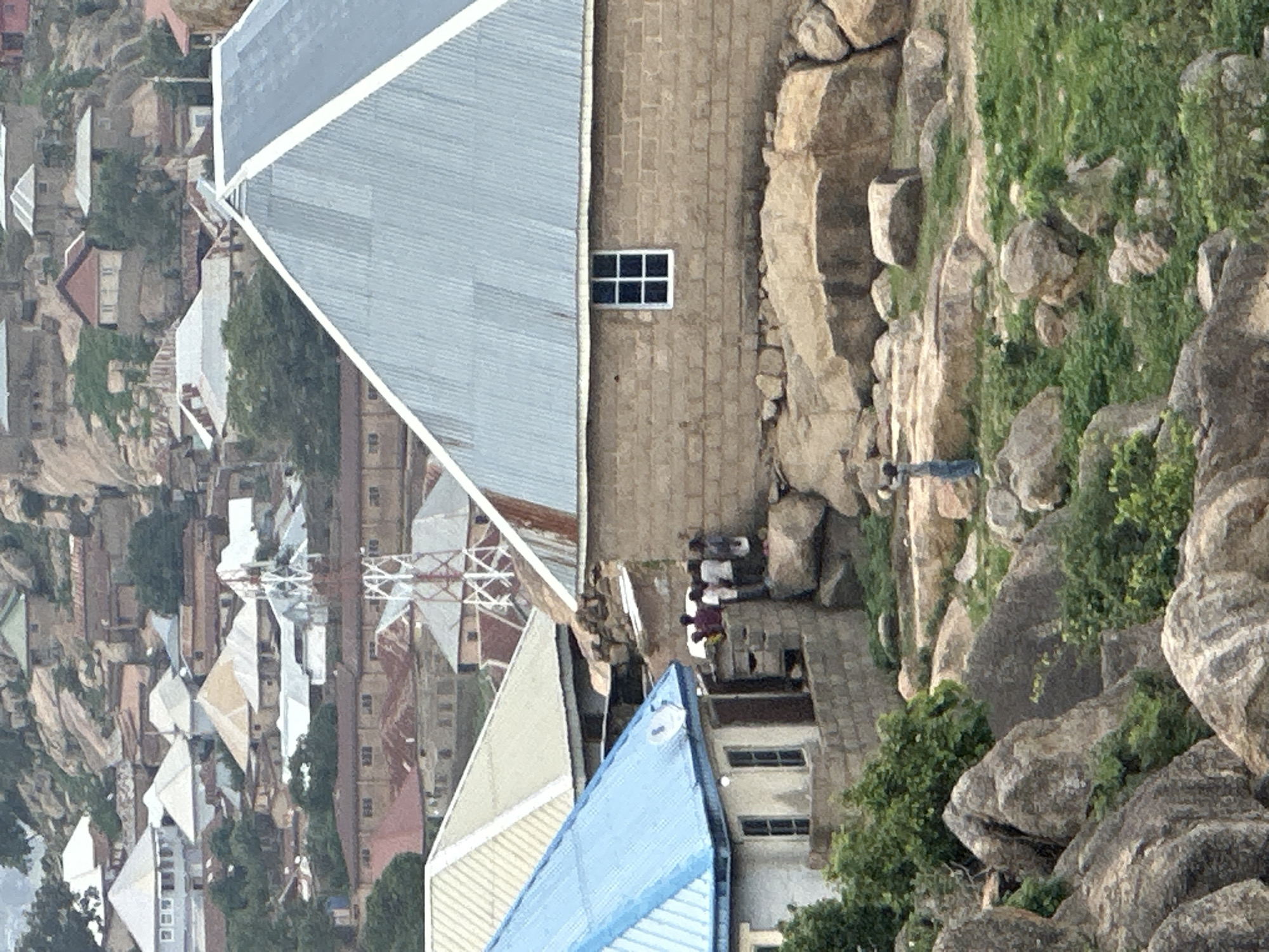 The image, presented on its side, depicts a dense settlement nestled amidst prominent rocky formations, consistent with a hilly region in Jos, Nigeria.
In the foreground (the right side of the image as presented), large, weathered boulders and sparse green vegetation define a steep, natural slope. The midground features several buildings integrated with the rocky landscape. A prominent structure has light brown brick walls and a large, angled grey corrugated metal roof, with a small, white-framed window with black panes visible on one wall.
A group of approximately three to four people are gathered on a dirt or rocky path below this building, appearing to be standing or resting. Further along the path, a solitary individual, dressed in a dark top and light trousers, stands looking towards the background. Another building with a striking blue corrugated roof is visible in the lower central part of the image.
The background (the left side of the image as presented) reveals a sprawling residential area. Numerous houses with various light-colored roofs (including white, off-white, and light brown corrugated iron, and some reddish-brown tiles) are tightly packed, interspersed with occasional green trees. A distinct lattice tower, likely for communication, rises above the distant buildings.
The scene is captured in bright, clear daylight, suggesting fair weather. There is no visible text. The overall impression is of a community thriving within a challenging yet scenic natural environment.