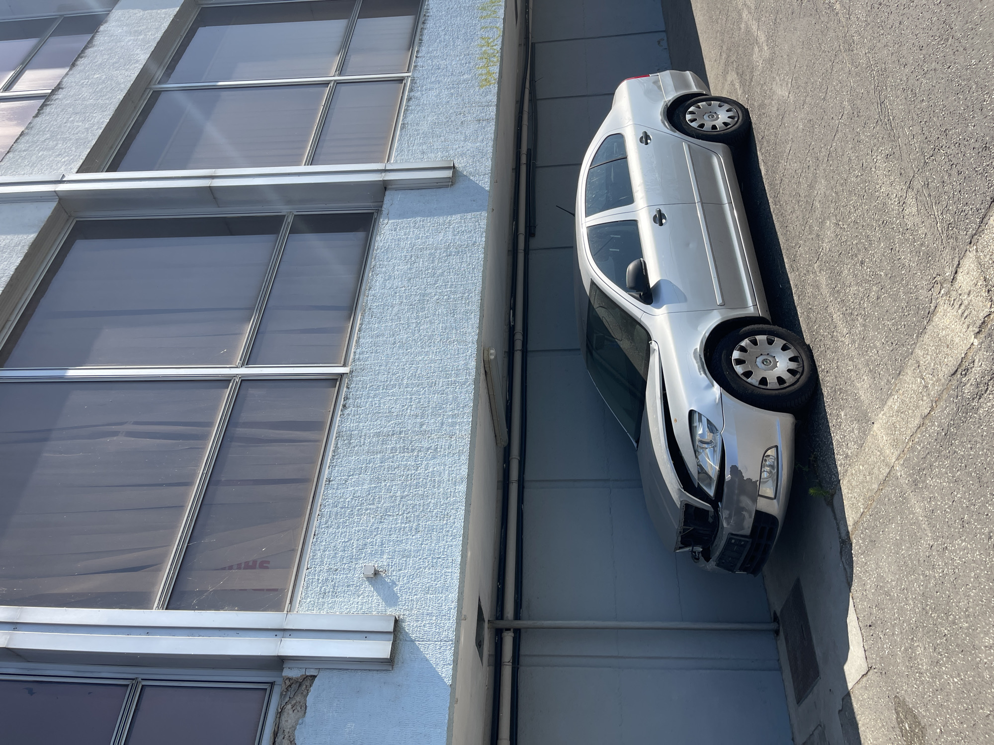 A silver sedan with a damaged front end is parked in front of a blue and gray building. The car's hood is crumpled and the front bumper appears to be damaged. The building has large windows with vertical blinds and a textured blue facade on the lower level. The parking area is paved with asphalt and concrete. The scene appears to be in an urban environment, possibly an industrial or commercial area. There is no graffiti visible on the building in the provided crops. The image is captured from a slightly low angle, looking up towards the building and the car. Sunlight is casting shadows, indicating a clear day. The overall impression is one of neglect or disuse, suggested by the damaged vehicle and the somewhat weathered appearance of the building.