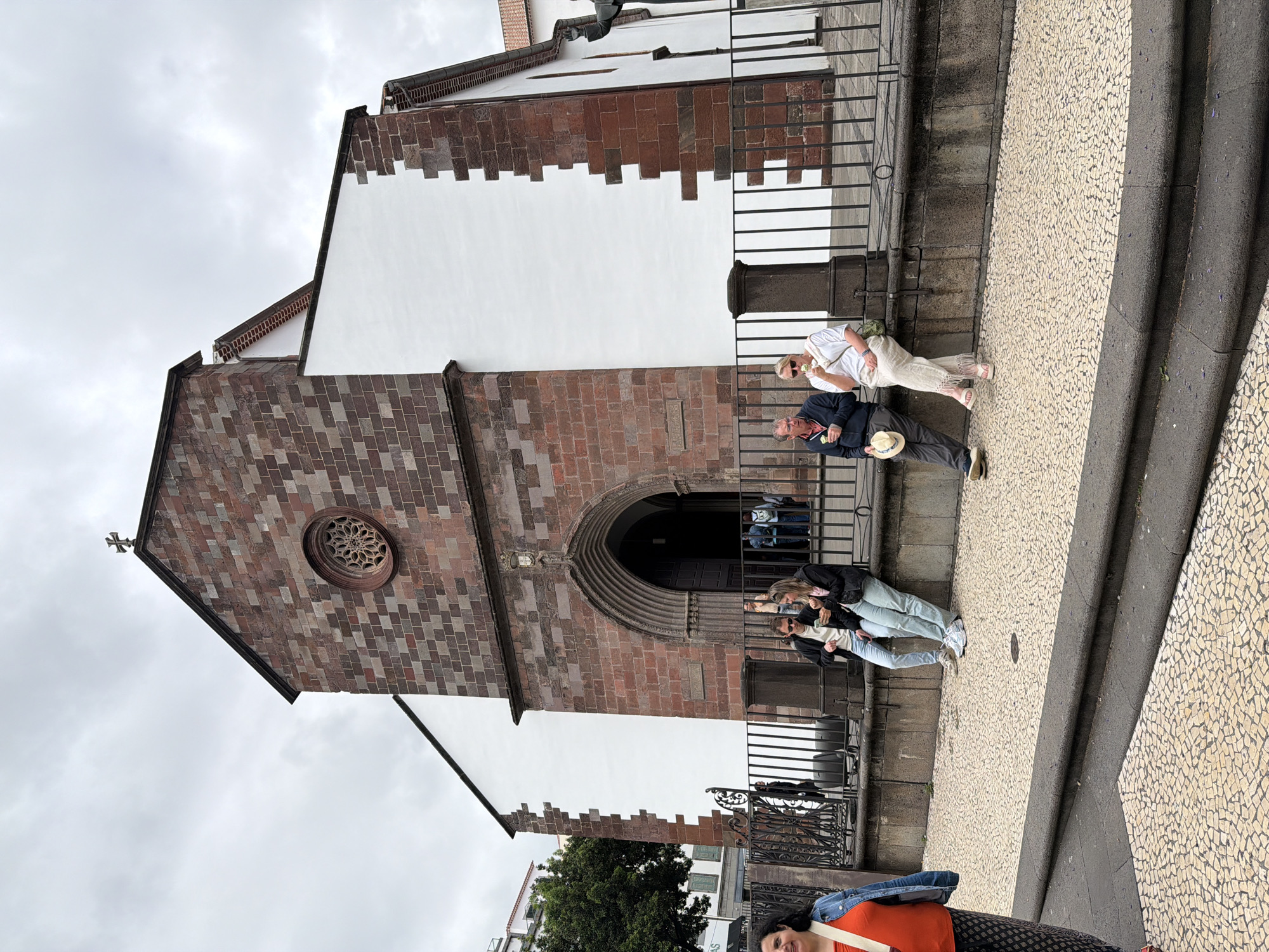 The image depicts a view of the Igreja do Colégio (Church of the College) in Funchal, Portugal. The church's facade is characterized by alternating panels of white stucco and reddish-brown brickwork. A large arched entrance is visible, as is a circular window above the door. The building is situated on a paved street composed of small, patterned tiles. Several tourists are present in front of the church, including people standing near a black metal railing and others walking along the sidewalk. The weather appears to be overcast.