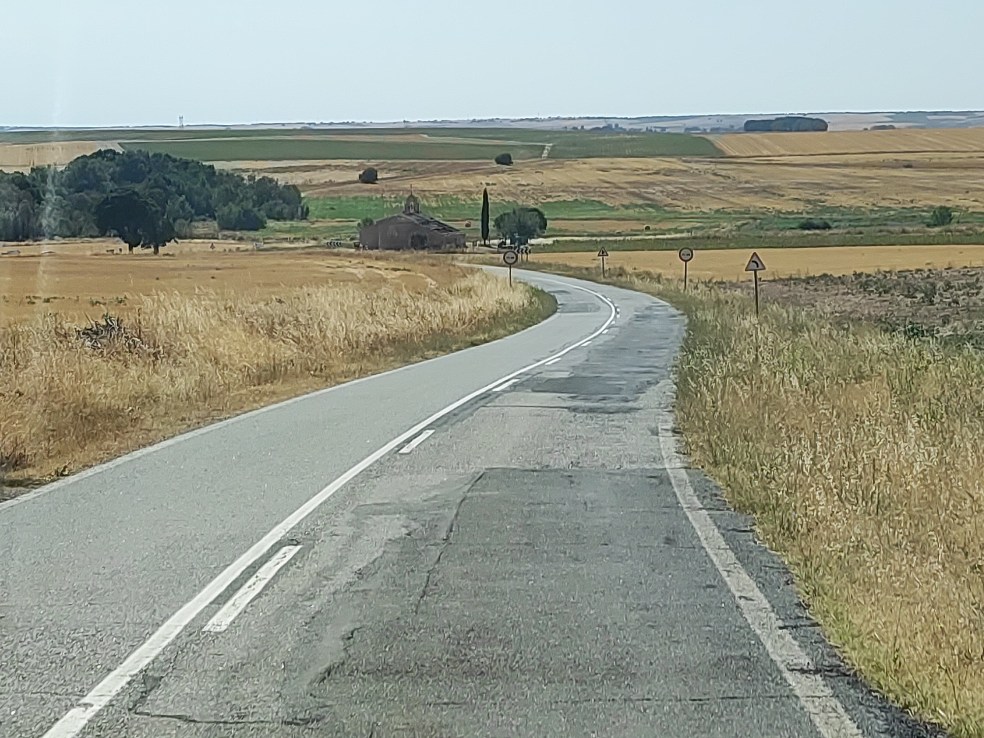 The image depicts a road curving through a rural landscape. The road surface is asphalt with clear white lane markings and appears to be in fair condition with some patching visible. On the left side of the road, a field of dry, golden grass and crops is present, bordered by a cluster of green trees in the middle distance. Further in the distance, on the left, is a large, dark green tree.
On the right side of the road, a similar field of dry grass and vegetation extends to the horizon. Several road signs are visible. Approaching the curve, there are two circular signs, one with a red border and a black symbol that is difficult to discern due to resolution, and another with a red border and a number "30" inside. Further along the curve, a triangular warning sign with a black symbol indicating a sharp turn to the left is visible.
In the mid-ground, nestled amongst the fields, is a small, stone building that appears to be a church, with a domed roof and a cross on top. A tall, slender tree stands next to the church. The landscape beyond the church is a patchwork of agricultural fields, with varying shades of green and golden-brown, suggesting different crops or stages of growth. The horizon is a flat line under a pale, hazy sky. The overall impression is one of a quiet, sunny day in a rural, agricultural region of Spain, possibly near Santa María la Real de Nieva, given the architectural style of the church and the characteristic landscape. No people are visible in the scene.