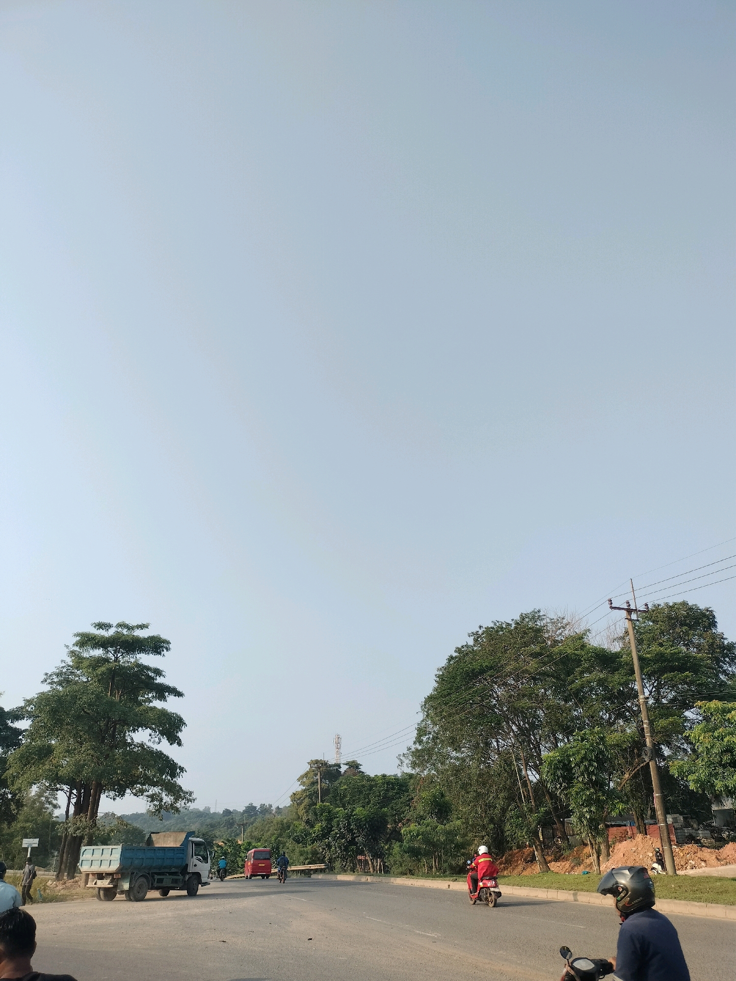 The image depicts a road scene in Batam, Indonesia. A blue dump truck is parked on the side of the road. Further down the road, a red van and two individuals on motorcycles are visible. The sky is clear and blue. Trees line the side of the road, and there are some construction materials scattered on the ground. The overall impression is of a typical day in a developing area of the city.