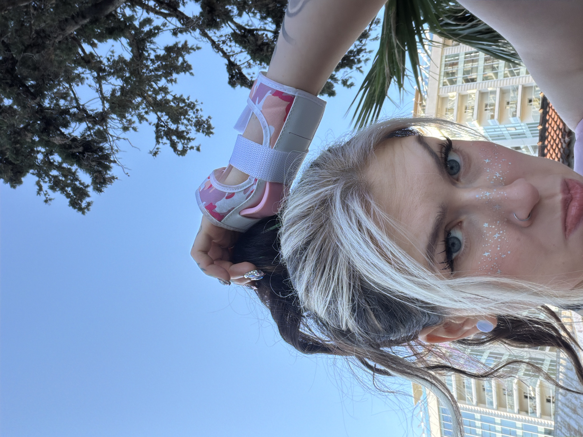 A low-angle selfie taken in Budva, Montenegro shows a fair-skinned woman with light blue eyes and glitter star makeup on her face. She is wearing a pink-and-white wrist brace on her left arm and a light blue stud earring in her left ear. Her dark brown hair has a blonde streak near her forehead and is partially tied up. Her nails are decorated with silver rhinestones. A partial view of a building with many windows is visible in the background, along with some palm tree fronds. A tree with dark green leaves is also visible in the upper left corner of the frame. The sky is clear and light blue.