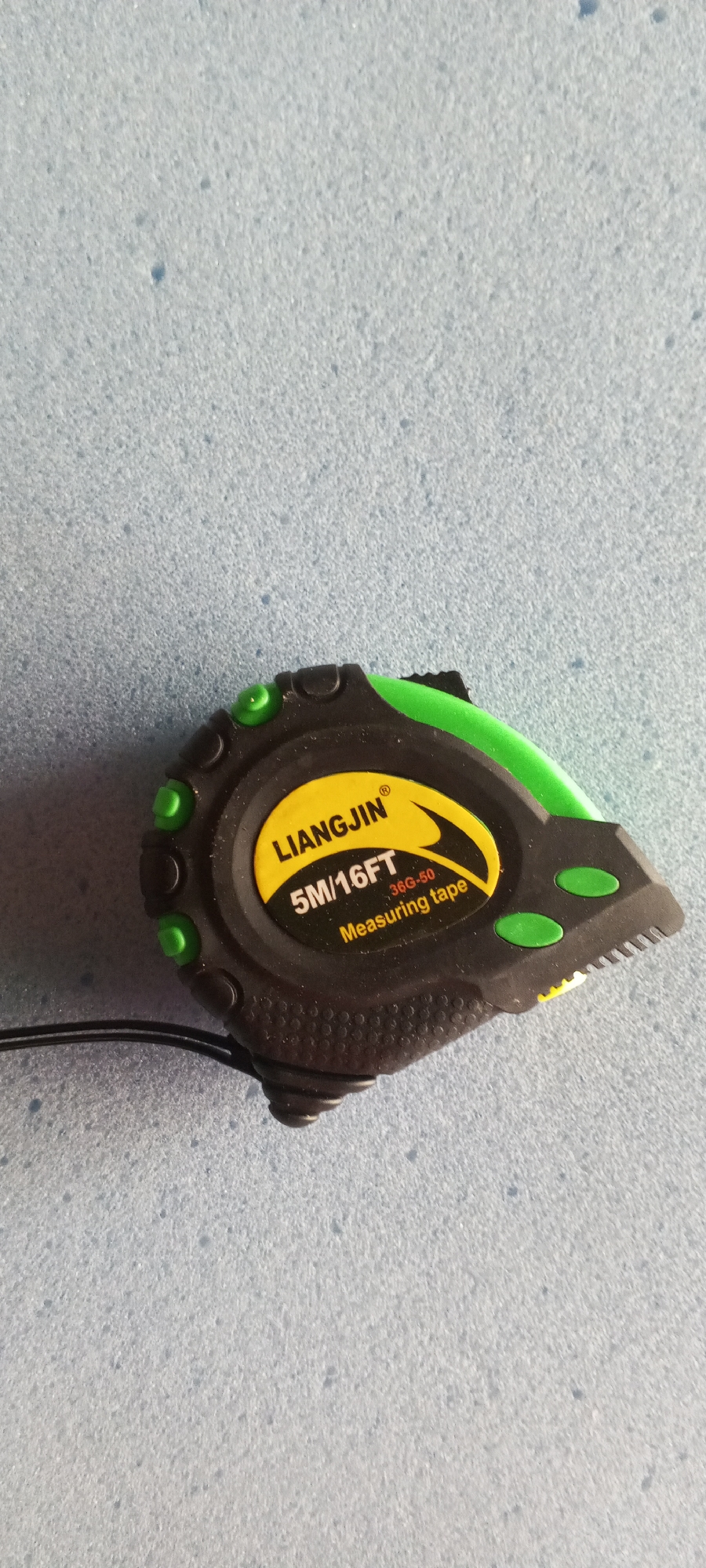 A black and green measuring tape is lying on a light blue, textured surface. The measuring tape has yellow labeling that reads "LIANGJIN 5M/16FT 36G-50 Measuring tape." The tape is partially extended and the end of the tape has a serrated edge. There is a black cord attached to the measuring tape. The textured surface appears to be a mat or a rug. There are no people or any indication of the city of Jalingo, Nigeria in the image.