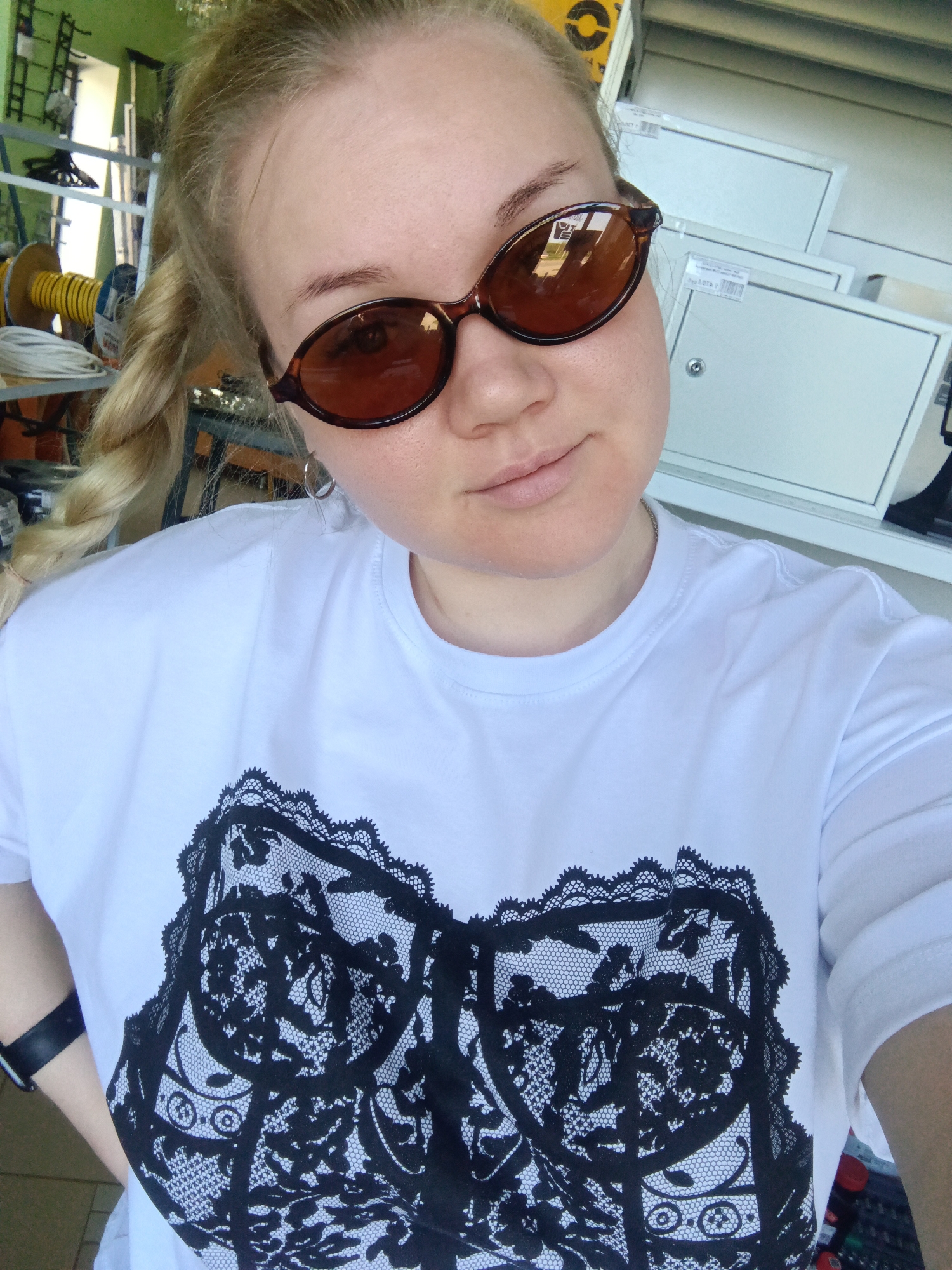 A selfie captures a fair-skinned woman with blonde hair styled in a side braid, adorned with brown oval sunglasses. She is wearing a white t-shirt featuring a black lace design on the front. A black wristwatch is visible on her left wrist. The background is slightly cluttered, containing coils of wire and stacked metal cabinets. The location is not discernible in the city of Фоки, Russia. The scene is well-lit and appears to be indoors.