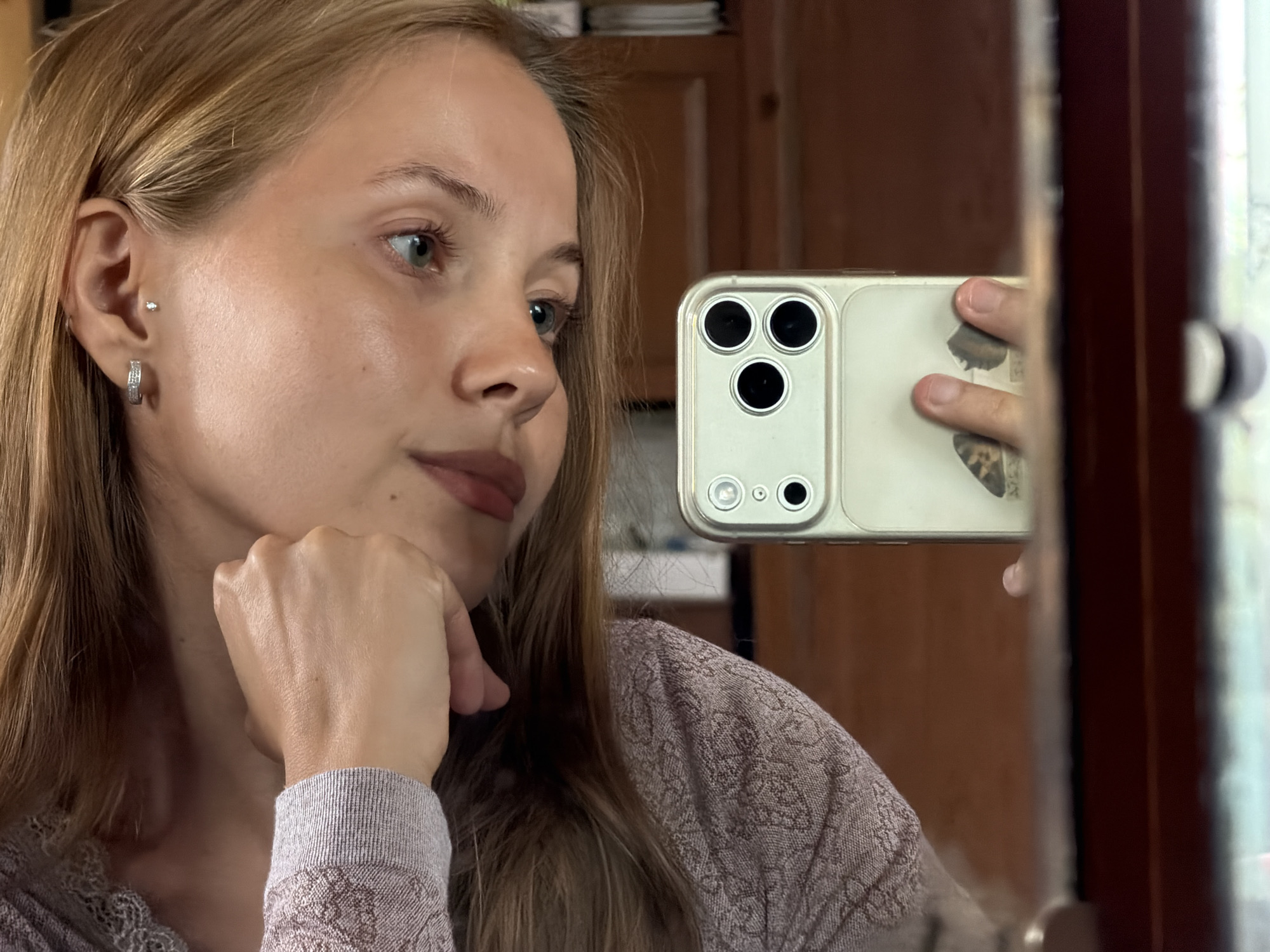 A young woman with blonde hair and blue eyes is taking a selfie in a mirror. She is wearing a light purple sweater with a lace trim and a patterned design. She has a small stud earring and a hoop earring in her left ear. Her right hand is resting on her chin, and she is looking off to the side. In her left hand, she is holding a smartphone, which is angled to capture her reflection. The smartphone case is light-colored and has a triple camera setup. The background appears to be an indoor setting, with wooden furniture and a window visible in the reflection. The lighting suggests it might be daytime.