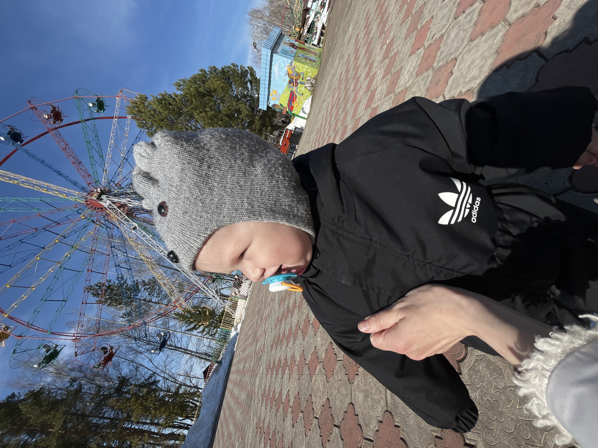 A toddler wearing a gray hat and black Adidas jacket is held by an adult, standing on a patterned brick pathway in Chaikovsky, Russia. In the background is a Ferris wheel, trees, and a small painted building. The sky is blue.