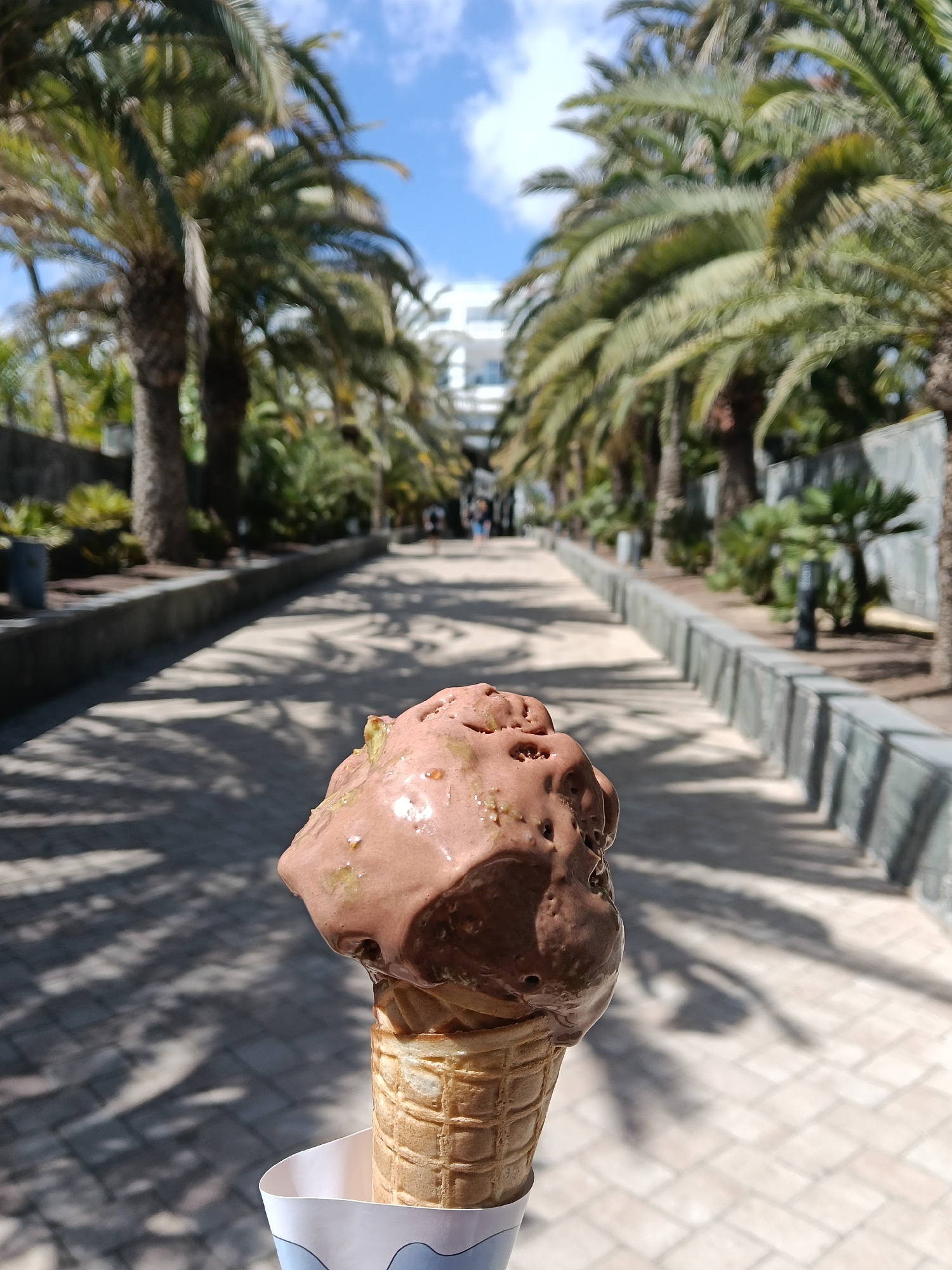 A chocolate ice cream cone is held in the foreground against a pathway lined with palm trees. The pathway, constructed of light gray pavers, stretches towards a white building in the distance. Shadows from the palm trees dapple the ground. People are seen walking in the distance, and the sky is blue with scattered clouds. The ice cream appears to be melting. The cone is held in a paper wrapper. The location is likely in San Bartolomé de Tirajana, Spain, given the prevalence of palm trees and the building style. Low concrete walls border the pathway on both sides.