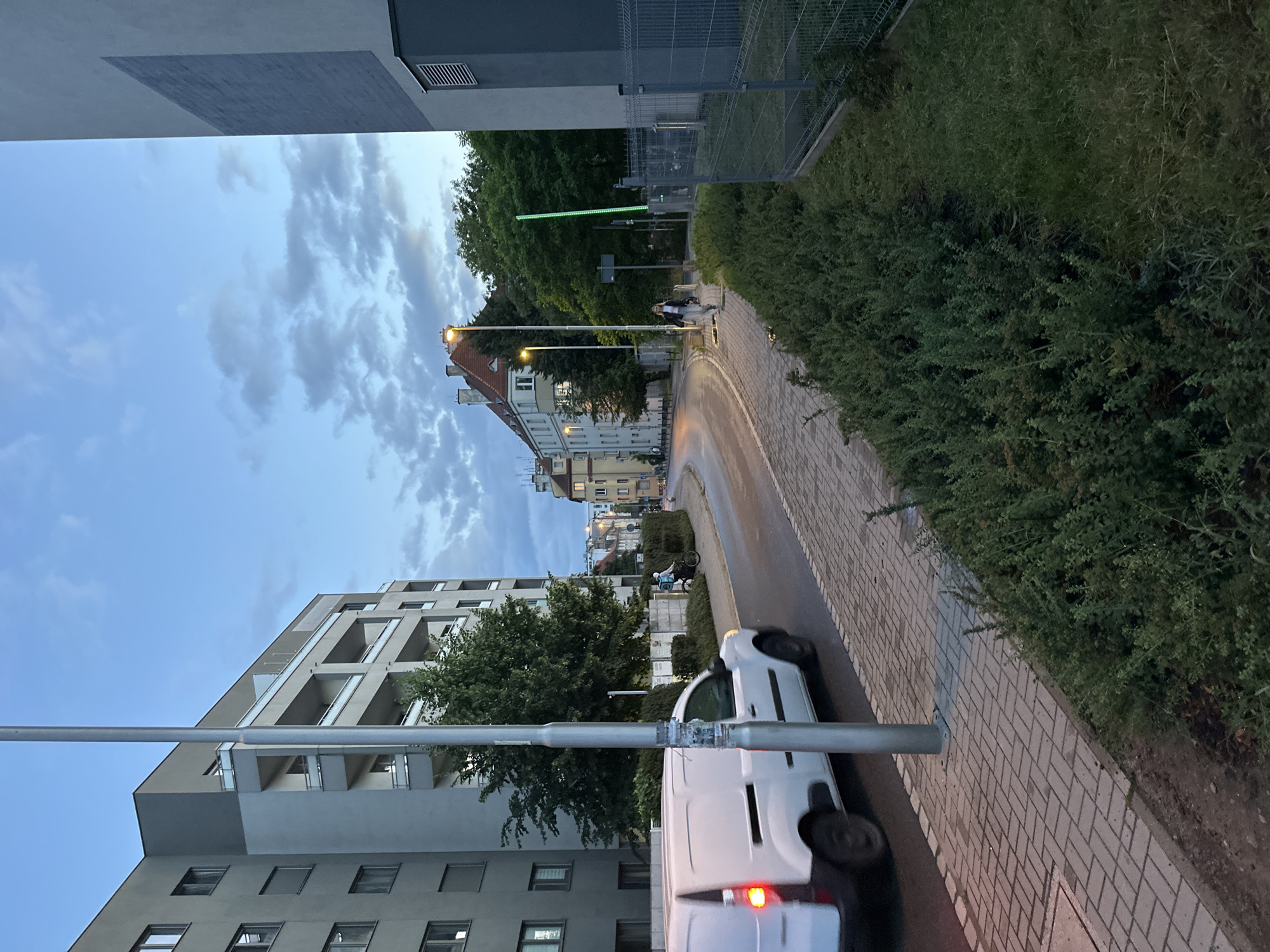 The image depicts a street scene in Wrocław, Poland, during twilight. A white van is in motion on the left side of the paved road, its brake lights illuminated. A lamppost stands prominently in the foreground, and a pedestrian walkway with a brick pattern lines the street. To the left of the walkway is a modern, multi-story building with balconies. Across the street, there are older, European-style buildings with red roofs, and a few trees. Further down the street, a person on a bicycle is visible, as well as another pedestrian walking on the sidewalk. The sky is a dusky blue with scattered clouds. Green bushes and grass are present on the right side of the image, adjacent to a fence.
