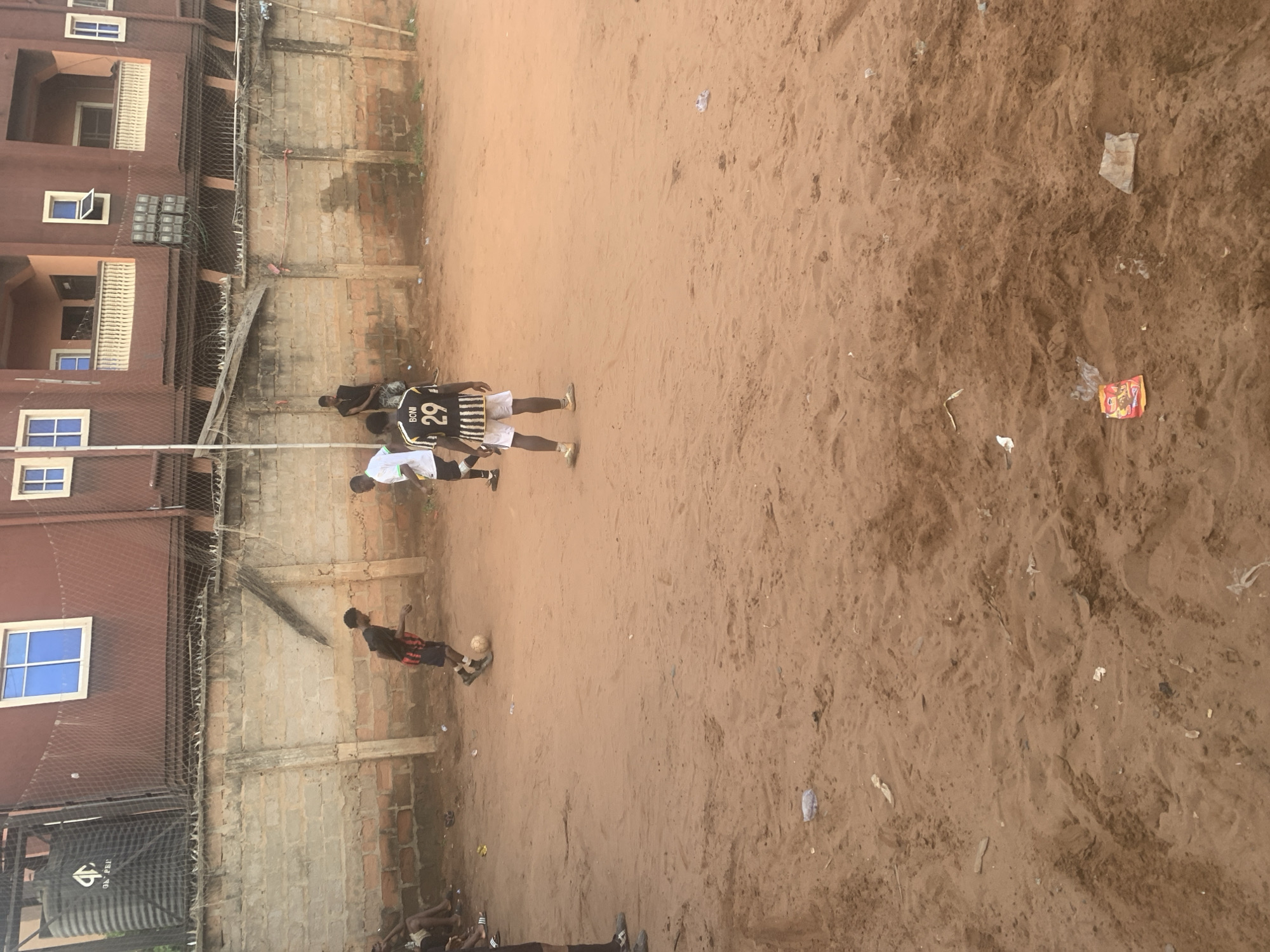 The image shows a dirt field in Onitsha, Nigeria. There are 5 people visible in the image. Three people are in the middle of the field. One person is wearing a black and white striped shirt with the number 29 on the back. One person is wearing white. One person is closer to the front. There is a building adjacent to the field. A water tank is visible on the property. Trash is scattered around the field. There is a soccer ball on the field.