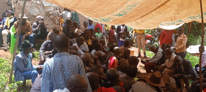 The image depicts a crowded outdoor gathering in Kundu 1, Democratic Republic of the Congo. Approximately 50 to 70 individuals, primarily adult males, are assembled under a large, light-colored tarp or canopy, possibly made from woven material with green markings, supported by rough wooden poles. Most attendees are seated directly on the reddish-brown earth ground, with some occupying a green plastic chair on the far left or resting on low, unseen structures.
Key figures include a man in the left foreground wearing a light blue suit jacket, blue jeans, and a black bowler hat, holding a small brown bottle. Another man in the immediate foreground, with his back to the viewer, wears a black and white patterned shirt. Various men throughout the scene wear casual clothing, including t-shirts, button-down shirts, and trousers; several wear hats such as baseball caps and fedoras. A few women and children are also visible among the seated crowd.
In the background, more individuals are standing, including one person in a yellow top and light-colored pants on the right, and another in a red shirt further left. The crowd appears attentive, with many individuals facing generally towards the right, suggesting a common point of focus or speaker. The environment features sparse green vegetation and dense foliage in the background, with the canopy providing shade from direct sunlight. No specific aggressive actions or overt interactions are discernible beyond general congregating.