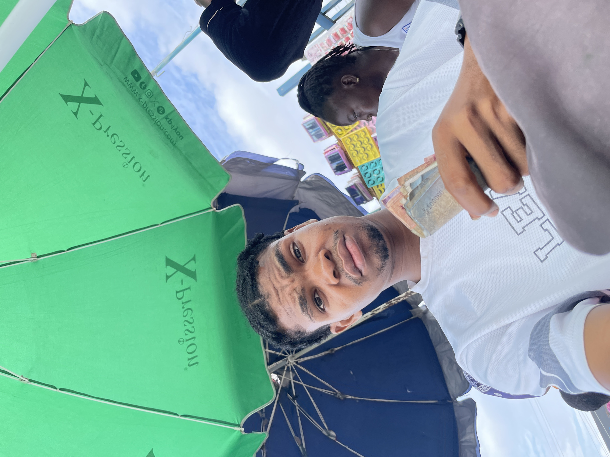 A young Nigerian man with braided hair is holding a fan of currency bills in his right hand, looking towards the camera. He is wearing a white t-shirt. He is seated under a large green umbrella, which is casting a shadow. To his right, a woman with braided hair is visible, wearing a white top. In the background, colorful amusement park rides are present, and the sky is cloudy. The scene appears to be in Ajah, Nigeria, potentially near a recreational area or market.