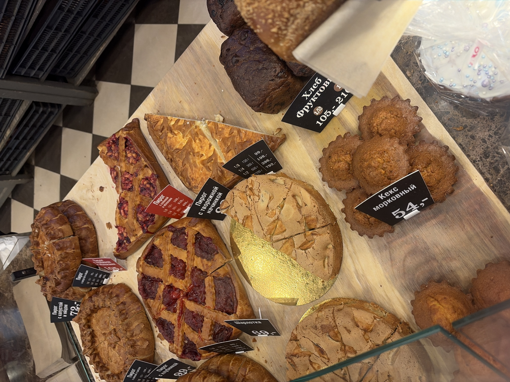 The image shows a variety of baked goods on display, presumably for sale. The items include different types of pies (with fillings like cabbage and egg, meat and rice, cranberry, apple, and a curd filling), a "Charlotte" cake, carrot cupcakes, and fruit bread. Price tags in Russian are visible next to each item, indicating the cost per fraction of the baked good (e.g., 1/8, 1/4, 1/2). The location is likely a bakery or a pastry shop in Saint Petersburg, Russia, as the labels are in Russian. A tray shelving unit can be seen on the left of the frame. The surface the baked goods rest on is made of light brown wood.