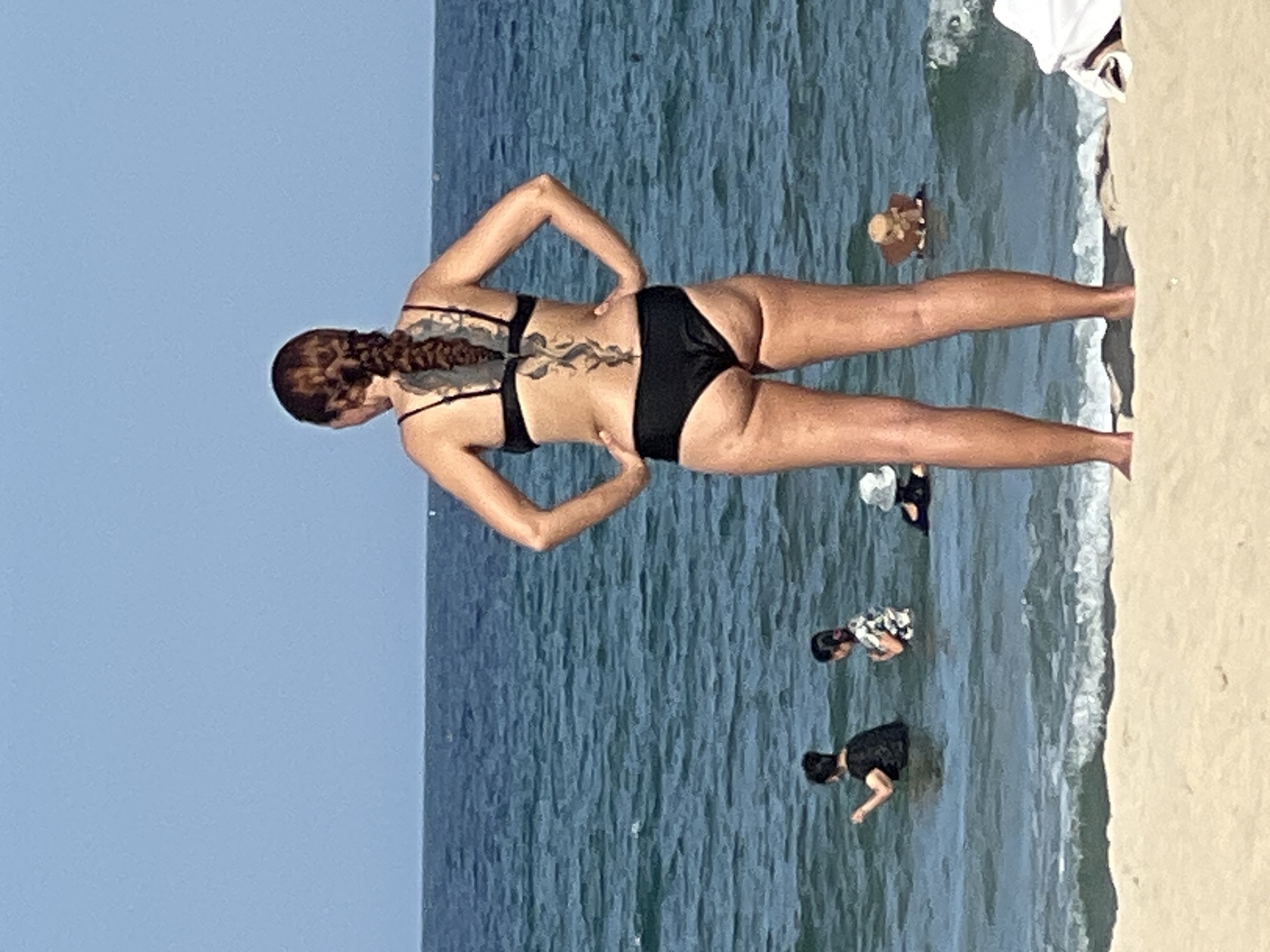In Đà Nẵng, Vietnam, on a bright, sunny day, a woman with auburn hair styled in a braid stands facing away from the camera on a sandy beach. She is wearing a black bikini and has a large, intricate tattoo extending down her spine. Her hands are on her hips. In the ocean behind her, several people are wading in the water. Two individuals, appearing to be younger, are closer to the shore, with one wearing a black top and the other a patterned top. Further out in the water, another person is visible wearing a white hat. In the distance, the vast expanse of the ocean meets the clear blue sky.