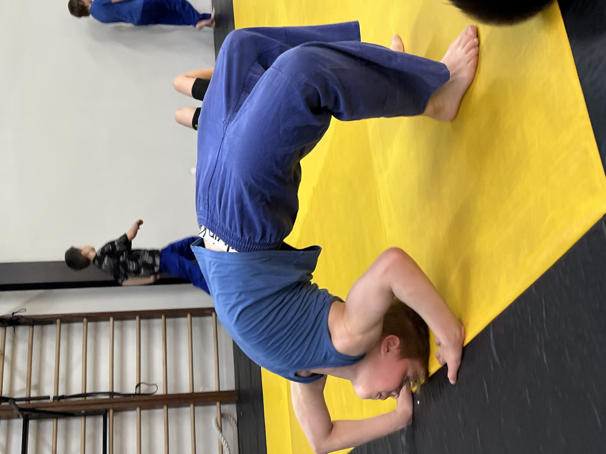 A youth dressed in blue training attire is doing a headstand on a yellow and black mat. The location is possibly a gym or training facility in Krasnoyarsk, Russia, given the surroundings, equipment, and training context. Another individual in similar clothing is visible in the background.