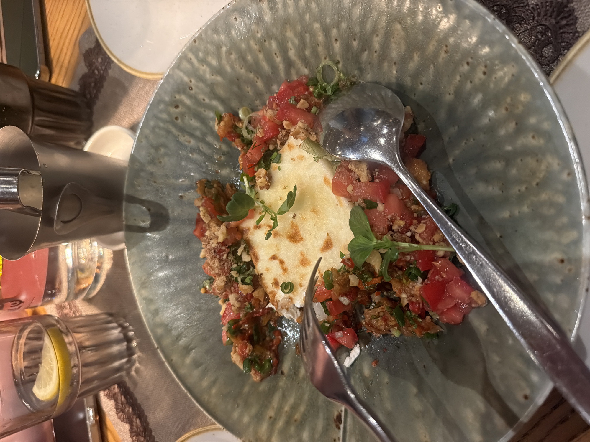 An overhead shot captures a dining scene in Sofia, Bulgaria, focusing on a savory dish served in a textured, greenish-grey ceramic bowl. The dish features a central, light-colored, possibly pan-fried or baked base (like cheese or flatbread) topped generously with diced red tomatoes, chopped green herbs (resembling scallions or chives), and a crumbly brown topping that could be nuts or toasted breadcrumbs. Fresh microgreens or small herb sprigs garnish the dish.
A metallic spoon rests on the top right side of the dish, partially submerged in the food, while a metallic fork is visible on the bottom right, also partially in the food, suggesting the meal is either being served or actively consumed.
To the left of the main dish, a clear glass contains water with a slice of lemon, indicative of a refreshing drink. Other glasses and a metallic jug or bottle are also visible on the table. The dining surface is a light-colored wooden table, partially covered by a patterned, dark beige or brown textile mat or coaster. A white plate with a gold rim is partially visible in the background.
The lighting suggests an indoor setting, likely a restaurant or a well-lit home, but the time of day, weather, and emotions of any individuals present are not discernible. There is no visible text in the image.