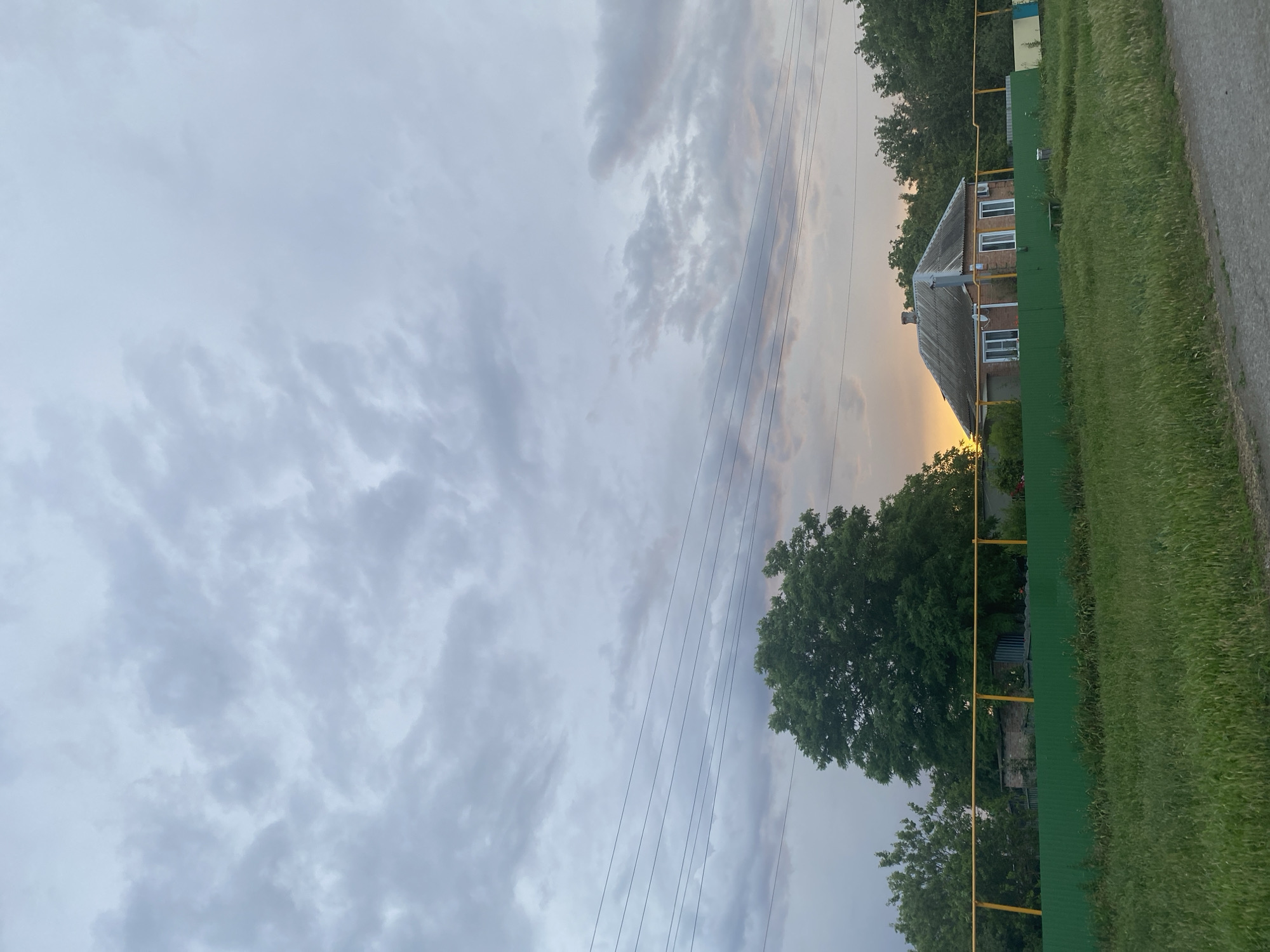 This is a vertical, medium-shot photo of a rural landscape at dusk in Ryasnoi, Russia. The scene is dominated by a dramatic sky filled with textured, grey clouds, suggesting an impending storm or the aftermath of one. A sliver of warm, orange light breaks through the clouds on the right side of the frame, indicating the setting sun.
In the mid-ground, a modest, brick house with a grey, sloped roof is visible. It has several white-framed windows. A tall, grey chimney rises from the roof. The house is bordered by a bright green fence and a yellow gas pipe runs along it. Behind the house, lush green trees with full canopies form a dense backdrop, partially obscuring other structures or foliage. To the right of the house, a strip of tall, green grass lines the edge of a grey asphalt road, suggesting the proximity of a rural road.
Several thin, black electrical wires stretch diagonally across the sky from the left to the right of the frame, hinting at the presence of a power line. No people are visible in the image. The overall mood is serene, perhaps a bit somber due to the cloudy sky, but with a touch of beauty from the setting sun's glow. The time of day is clearly evening, as indicated by the sunset colors and the general lighting.