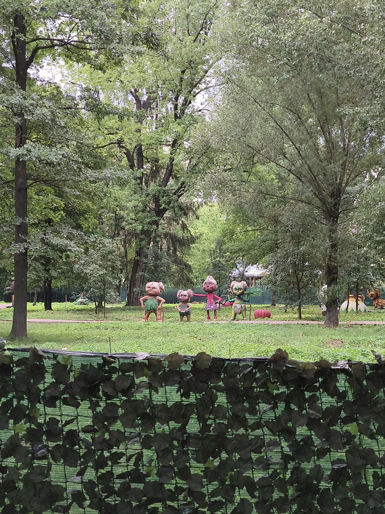In the city of Ternopil, Ukraine, a park displays a scene with several cartoonish figures in a grassy area surrounded by trees. In the foreground, a black mesh fence covered with artificial green leaves partially obscures the view. Behind the fence, on the left side of the park, a tall, dark tree stands prominently. In the middle ground, four figures are arranged in a line on a wooden path. The first figure on the left is a male character with brown skin and a green loincloth, holding a stick. Next to him is a smaller, child-like figure, also in a green loincloth. To their right stands a female figure with pink hair and a pink dress, holding a green leaf. The fourth figure is a character with green, spiky hair and a green outfit, also holding a green leaf. Further back, to the right of these figures, a large, yellow flower-like structure and a lion-like creature are visible. The background is filled with lush green trees, with a building partially visible behind the foliage. The ground is covered in short, green grass.
