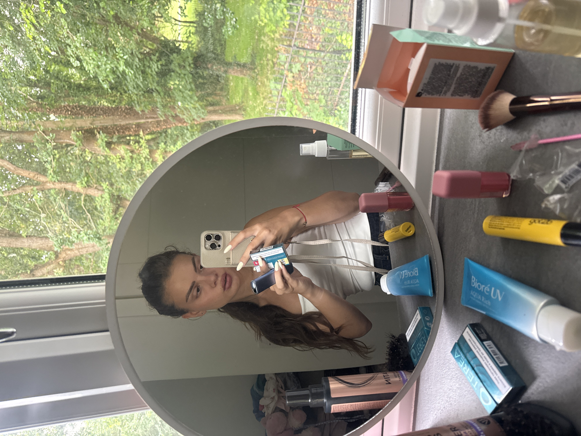 The image shows a young woman taking a selfie in a mirror, holding a phone and a package of what appears to be tobacco. She is standing in front of a window with trees and foliage visible outside, suggesting an outdoor, possibly rural setting. The area surrounding her, likely a bathroom or dressing area, is cluttered with various cosmetic products, including tubes of cream, makeup brushes, and bottles of sprays. One tube of cream is labeled "Biore UV Aqua Rich," indicating a skincare product. A hairspray bottle is also visible, along with a small pink lipstick or lip gloss. The overall scene suggests a moment of personal grooming or preparation, with the natural scenery outside providing a backdrop. There is no information to suggest this location is in Throop, United Kingdom; however, the presence of greenery and trees might be consistent with a rural or suburban area.