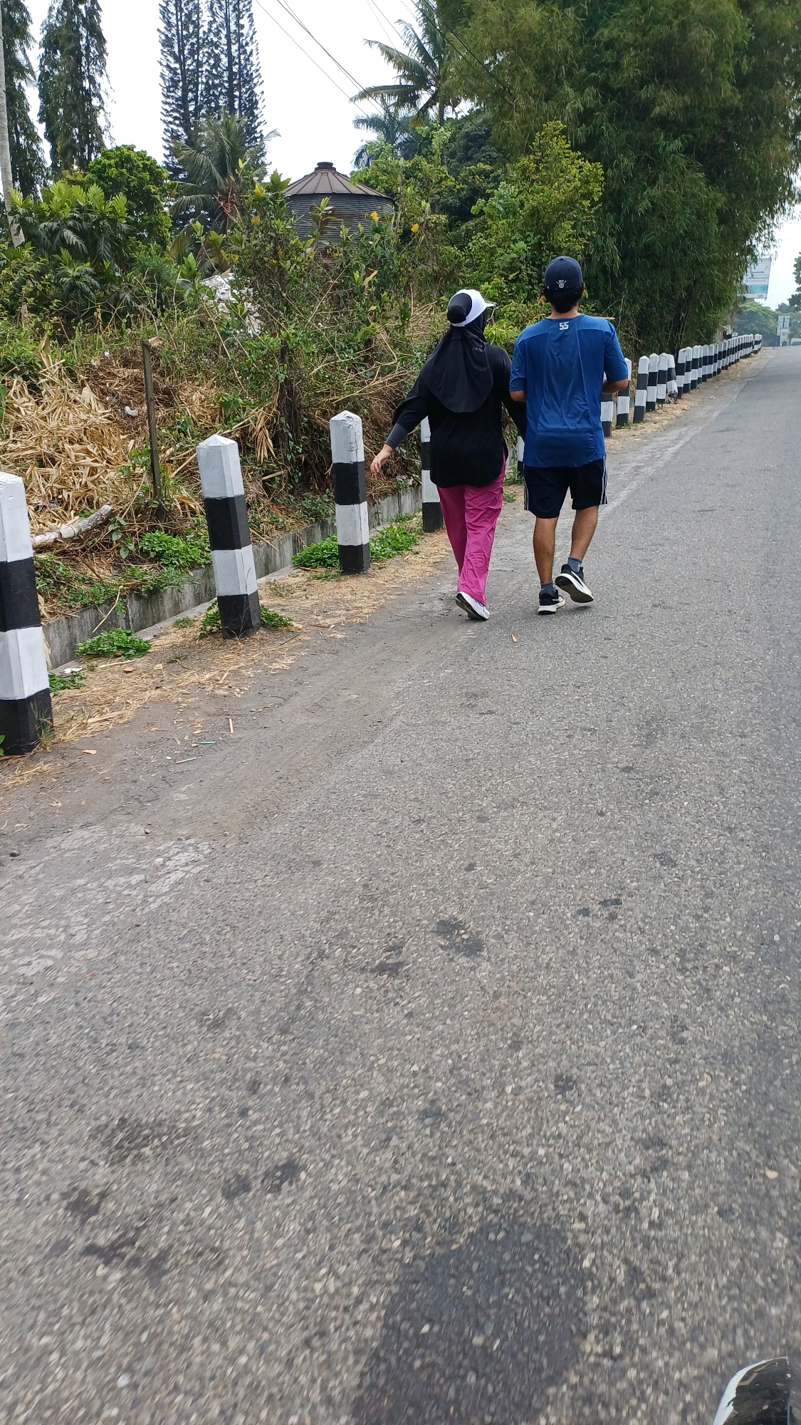 Two individuals, viewed from behind, are walking on an asphalt road bordered by a black and white striped barrier on the right and a dry, overgrown embankment with a concrete curb on the left. The individual on the left is wearing a black hijab and cap, and bright pink leggings. The individual on the right is wearing a blue t-shirt with the number "55" on the back and black athletic shorts. Trees and vegetation, including palm trees and bamboo, are visible in the background. A metal structure, possibly a water tank, is also present among the trees. The scene appears to be in a semi-rural or suburban area.