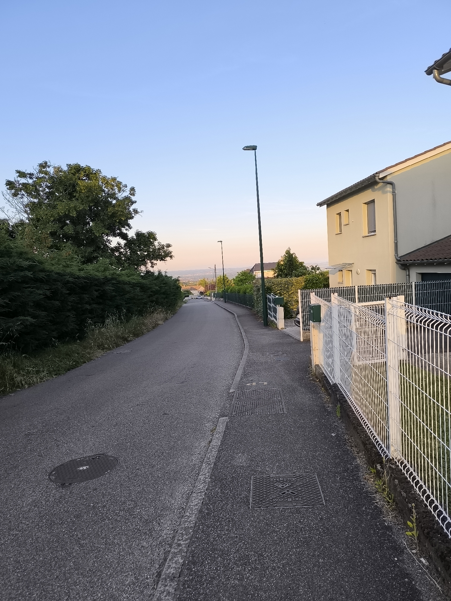 The image depicts a residential street in Pélussin, France. The street slopes downwards, flanked by houses on the right and dense greenery on the left. A white fence runs along the right side of the street, bordering a house with light-colored walls. Several utility access covers are visible on the pavement. Streetlights line the road. In the distance, a valley or plain is visible under a clear, slightly pink-tinted sky, suggesting early morning or late evening light. There are no people visible in the image.