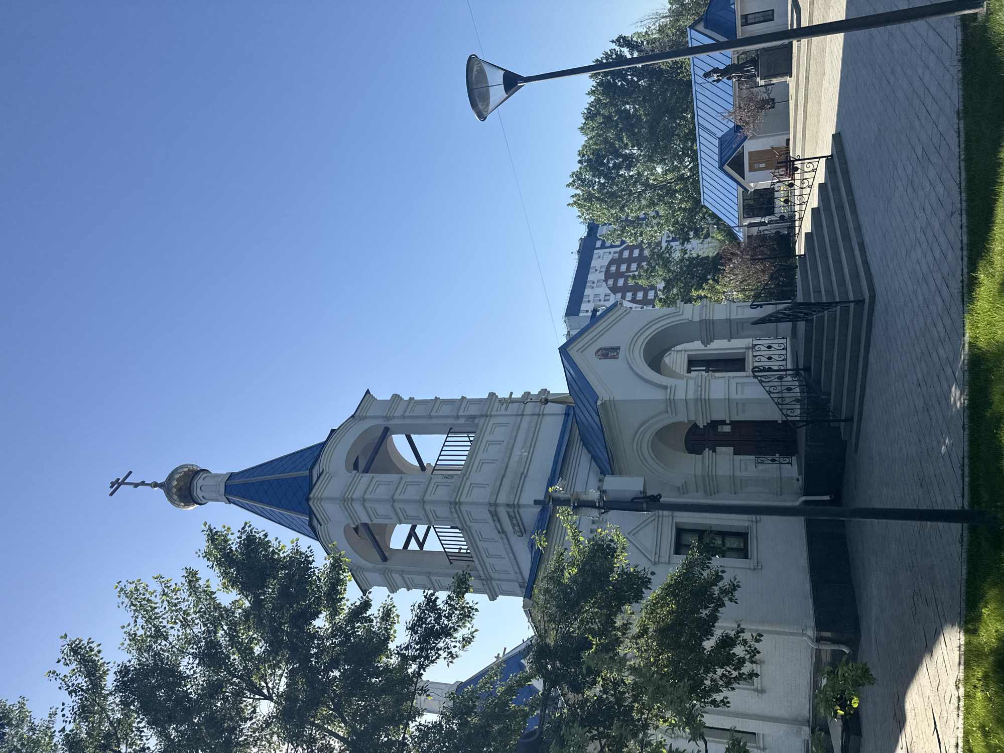 The image shows the Church of the Holy Great Martyr Catherine in the Vspolye in Moscow, Russia. The church is white with blue accents, including a blue roof and trim. There is a bell tower on the left side of the image. A cross tops the dome above the bell tower. The church is surrounded by trees and a paved walkway. A lamppost is located in the foreground, next to the church. A building is in the background to the left, and stairs leading up to another building are visible to the right, behind the church. The sky is blue.