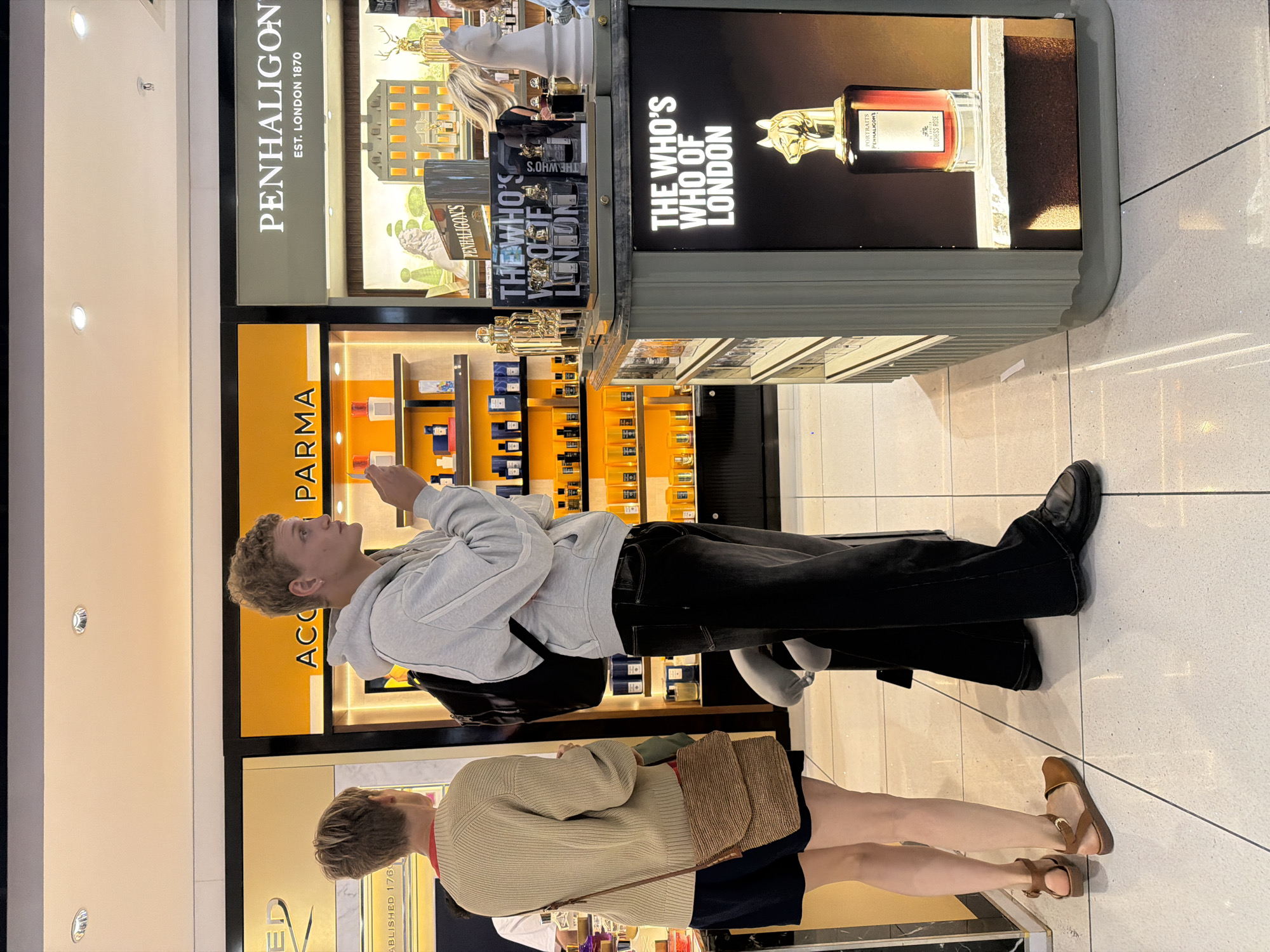 A young man with short, blond, curly hair is standing in an airport duty-free shop, holding a small white object to his nose. He is wearing a grey hooded sweatshirt, black jeans, and a black backpack. To his left, a person with short, blond hair, wearing a beige knitted sweater and dark shorts, is looking at a display of perfumes. The shop appears to be in Crawley, United Kingdom, and features prominent branding for "PENHALIGON'S EST. LONDON 1870" and "ACQUA DI PARMA." The floor is tiled with a light-colored pattern.