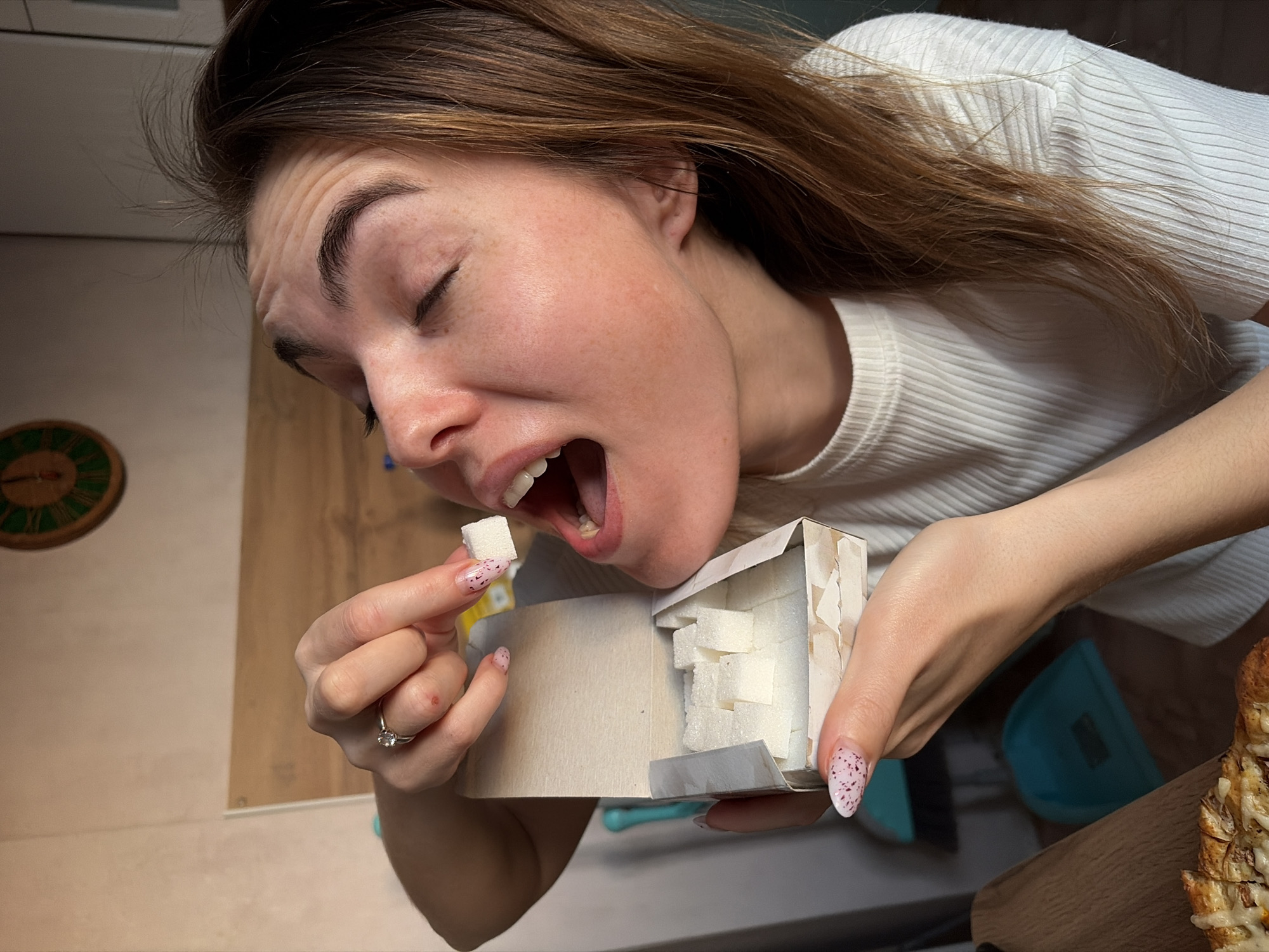 A woman is holding a box of sugar cubes with her right hand and a single sugar cube with her left hand, positioning the latter to put it into her open mouth. She is wearing a white ribbed shirt. Her hair is brown and shoulder-length. She is wearing a ring on her left ring finger and has manicured nails. The environment appears to be an indoor space, possibly a kitchen, with a wooden countertop and a clock on the wall. The exact location in Izhevsk, Russia, cannot be determined from this image alone.