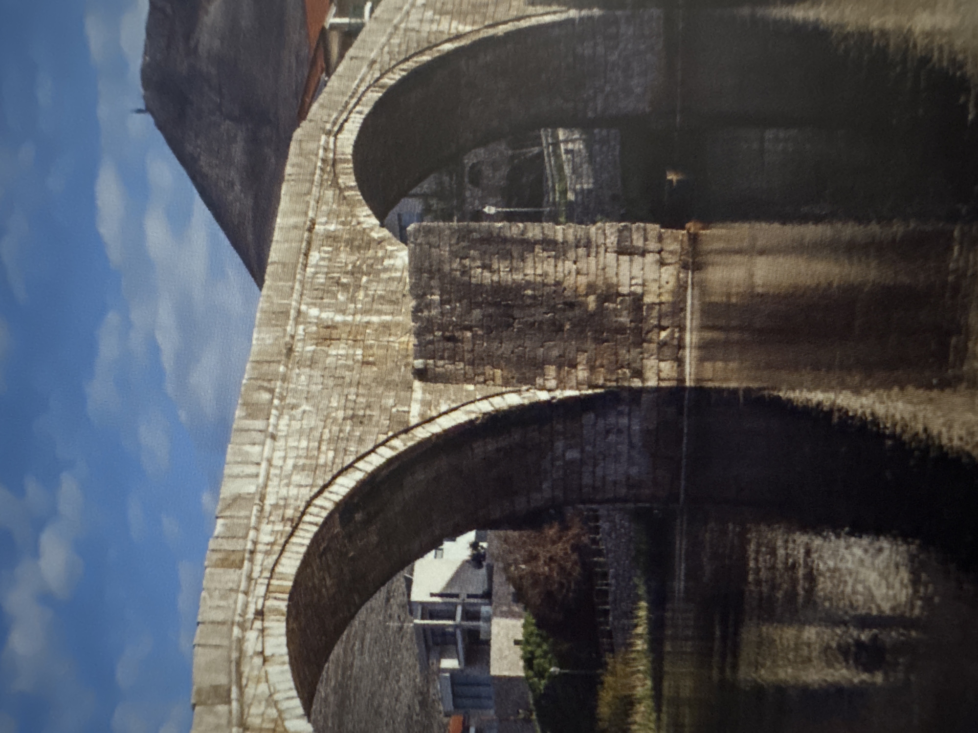 The image shows a stone arch bridge spanning a body of water, likely a river or a lake, in the city of Unknown, Unknown. The bridge is constructed from large, rough-hewn stone blocks, with two prominent arches visible. The water below reflects the bridge, creating a dark, shimmering surface.
To the left of the bridge, there are modern buildings with white walls and dark windows, and some vegetation. In the background, a large, dark hill rises, with a small human figure standing on its peak, suggesting a lookout point or a monument. The sky above is a clear blue with scattered white clouds, indicating a bright, possibly sunny day. The overall scene conveys a sense of history and permanence, with the ancient stone bridge contrasting with the modern buildings and the natural landscape.