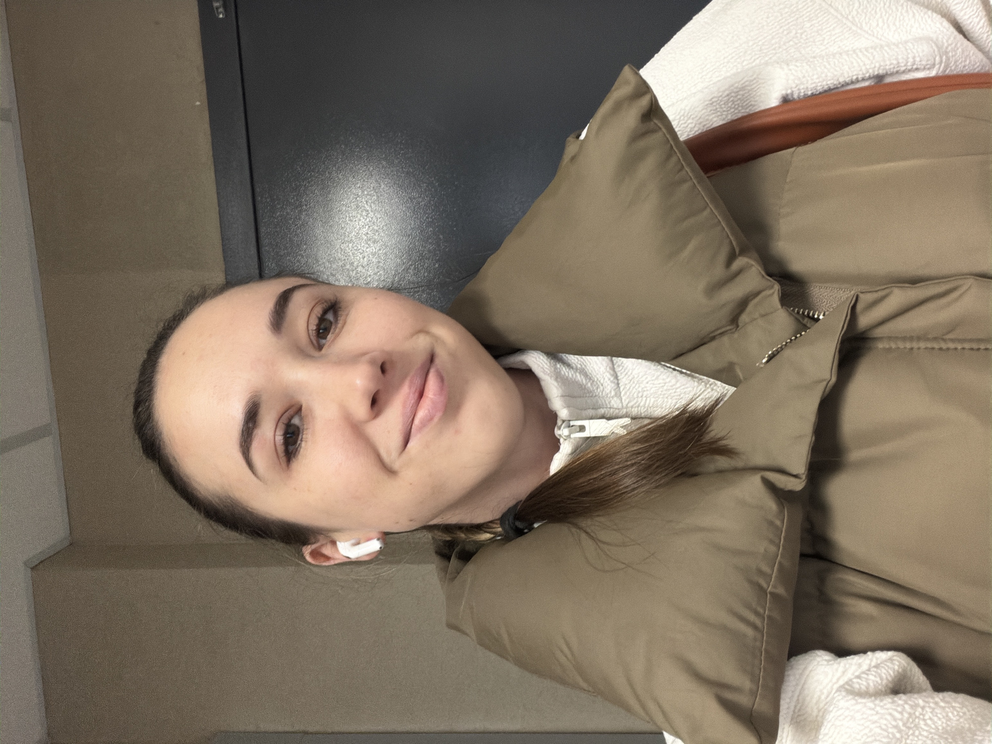 A young woman is pictured in a selfie. She has light skin, brown hair pulled back in a braid, and is wearing an earbud in her left ear. She is smiling slightly at the camera. She is wearing a puffy brown jacket over a white hoodie. The background consists of gray and black walls. The photo was not taken in Rostov-on-Don, Russia.