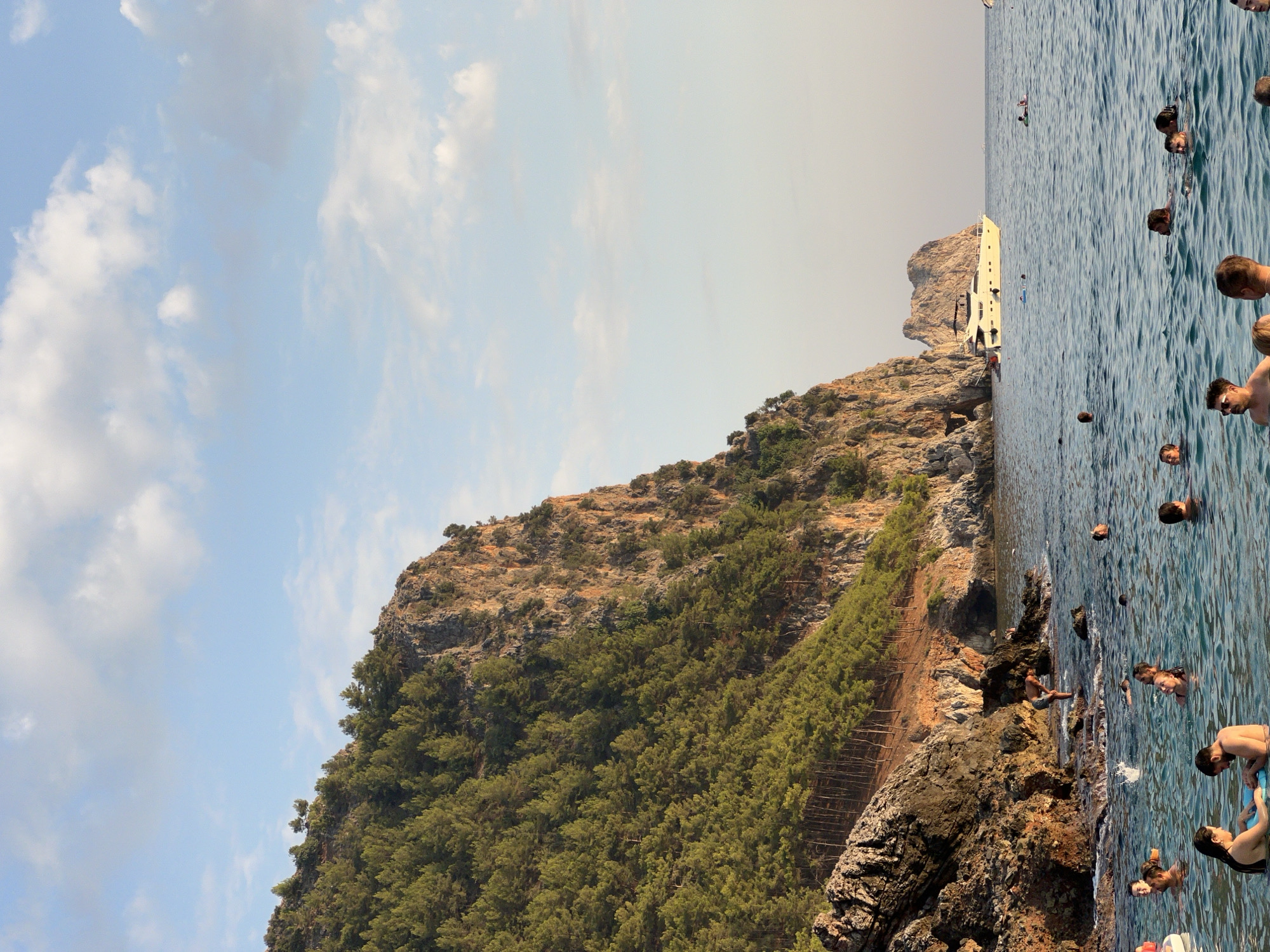 This is a photograph of a beach in Alanya, Turkey, with a rocky cliff face covered in trees dominating the left side of the frame. In the foreground, several people are swimming and wading in the clear blue water. A few individuals are using inflatable devices. In the middle ground, a white yacht is anchored offshore. The sky above is a light blue with scattered white clouds. The overall scene depicts a relaxed, summer atmosphere at a coastal location.