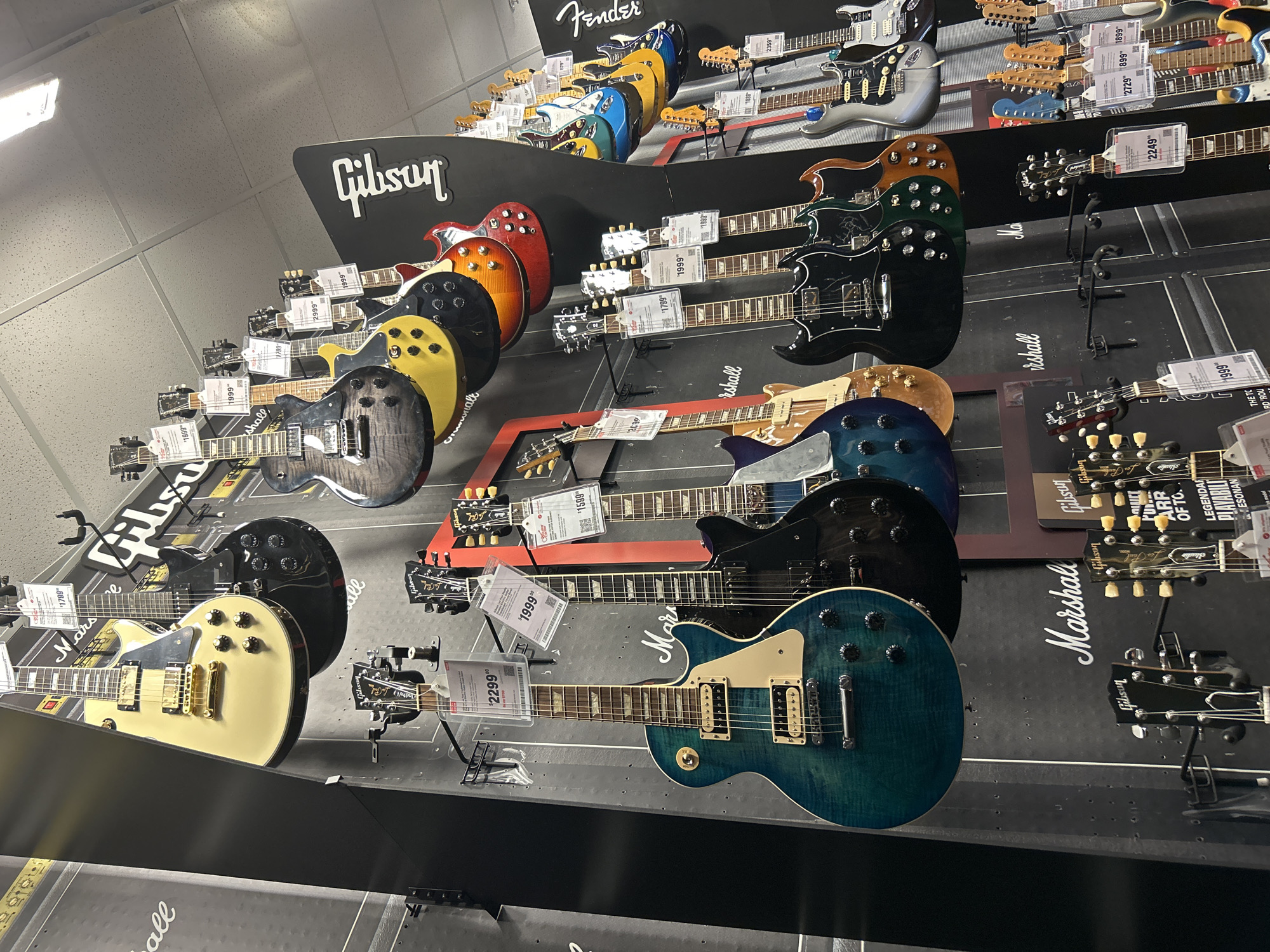 The photograph appears to be taken inside a music store. The main subjects are numerous electric guitars displayed on a wall. The guitars are various models and colors, including brands like Gibson and Fender, with price tags attached to some. The display wall is branded with the logos of Gibson and Marshall. The guitars are hanging on a black wall display. The prices of some of the guitars visible range from $1599 to $2999. Based on the presence of the "Fort Myers" tag and Gibson and Fender Guitars, this is likely a retail music store located in Fort Myers, Florida, USA.
