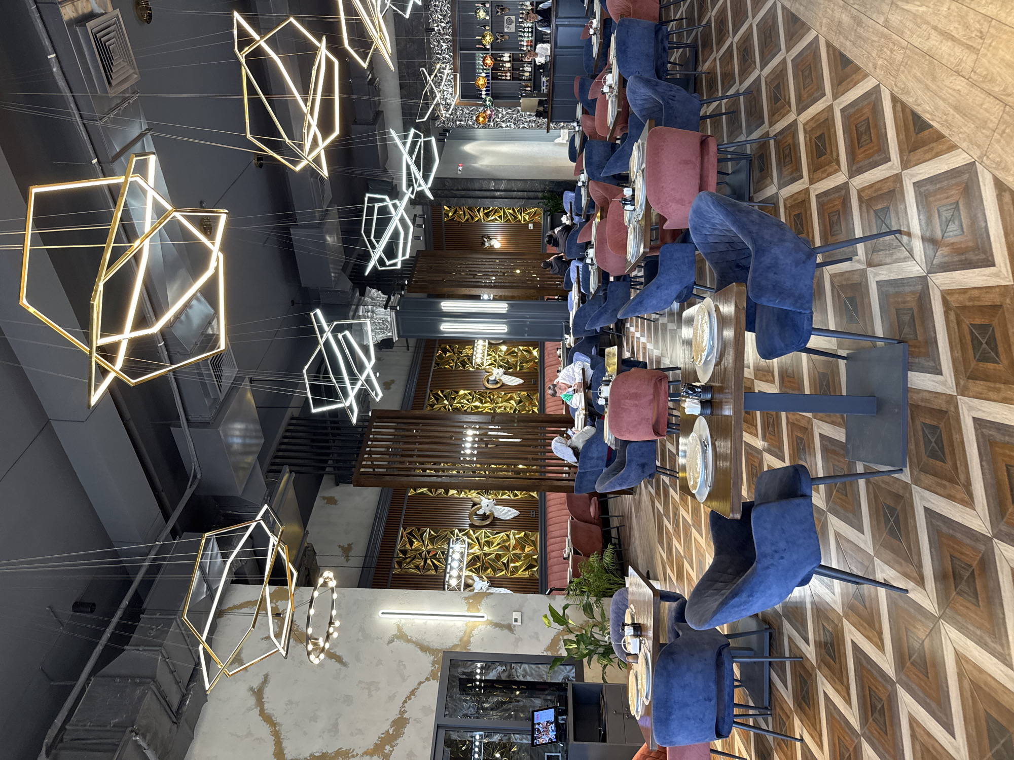 The image depicts the interior of a restaurant in Çolpon-Ata, Kyrgyzstan. The space is filled with tables and chairs arranged in rows. The chairs alternate between blue velvet and mauve fabric, and appear modern. The tables are set with plates and utensils.
The floor is made of wood tiles arranged in a geometric pattern. Overhead, there are several geometric chandeliers of gold and white light. The ceiling is dark and industrial.
Towards the back of the restaurant, a bar is visible. The walls are decorated with metallic geometric patterns and a horizontal line of white fluorescent lights. There are a few people seated at the tables, some of whom appear to be dining. Plants and other decorative elements are interspersed throughout the space.
